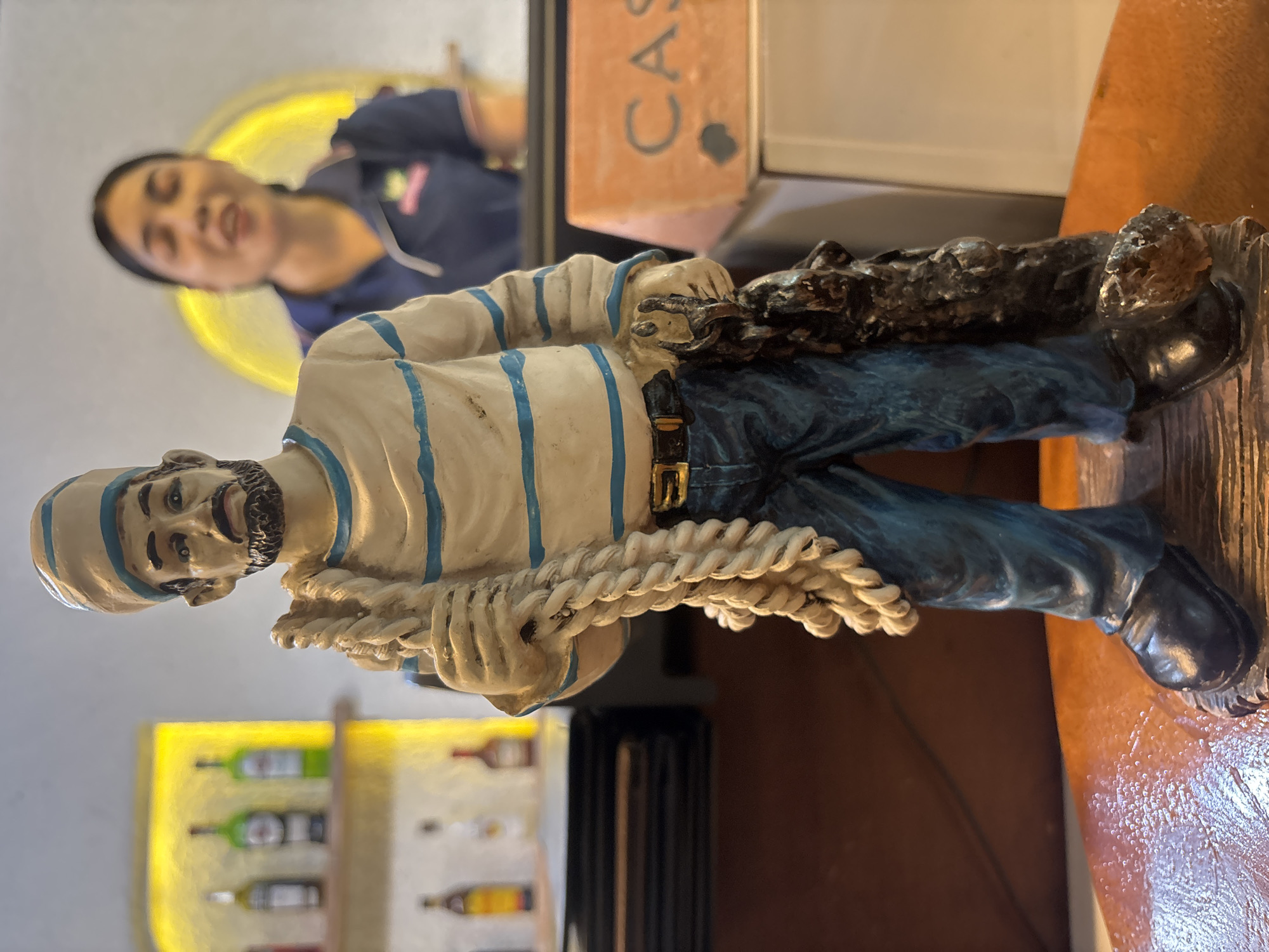 In Chalong, Thailand, a foreground figurine of a sailor stands resolutely. He is depicted wearing a striped shirt, blue pants, a belt, and a cap, and carries a thick rope over his shoulder and a dark, textured object in his right hand. Behind the figurine, a blurred woman in a dark blue polo shirt is visible, appearing to be speaking. The background reveals shelves stocked with bottles, suggesting a bar or shop setting. A sign with the letters "CAS" is partially visible to the right of the figurine.