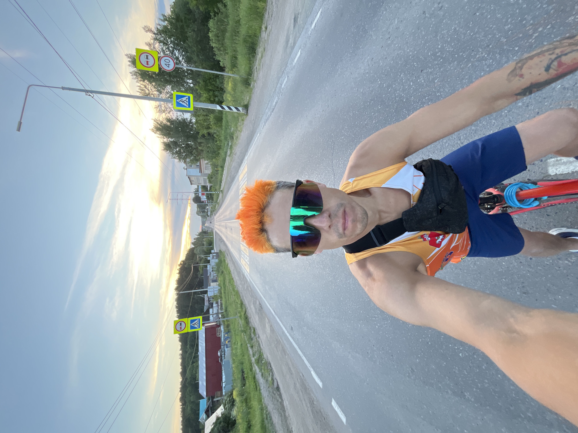A man with bright orange hair, wearing large, reflective sunglasses, an orange and white tank top, and blue shorts, rides a bicycle on a paved road. He has a black fanny pack around his waist. The road is lined with green grass and trees, and there are houses and buildings in the distance. Traffic signs are visible along the side of the road, including a pedestrian crossing sign and a speed limit sign. The sky is a pale blue with wispy clouds, and the sun appears to be setting. The overall impression is that of a rural or suburban road on a clear evening. The specific location is the city of Chaikovsky, Russia.
