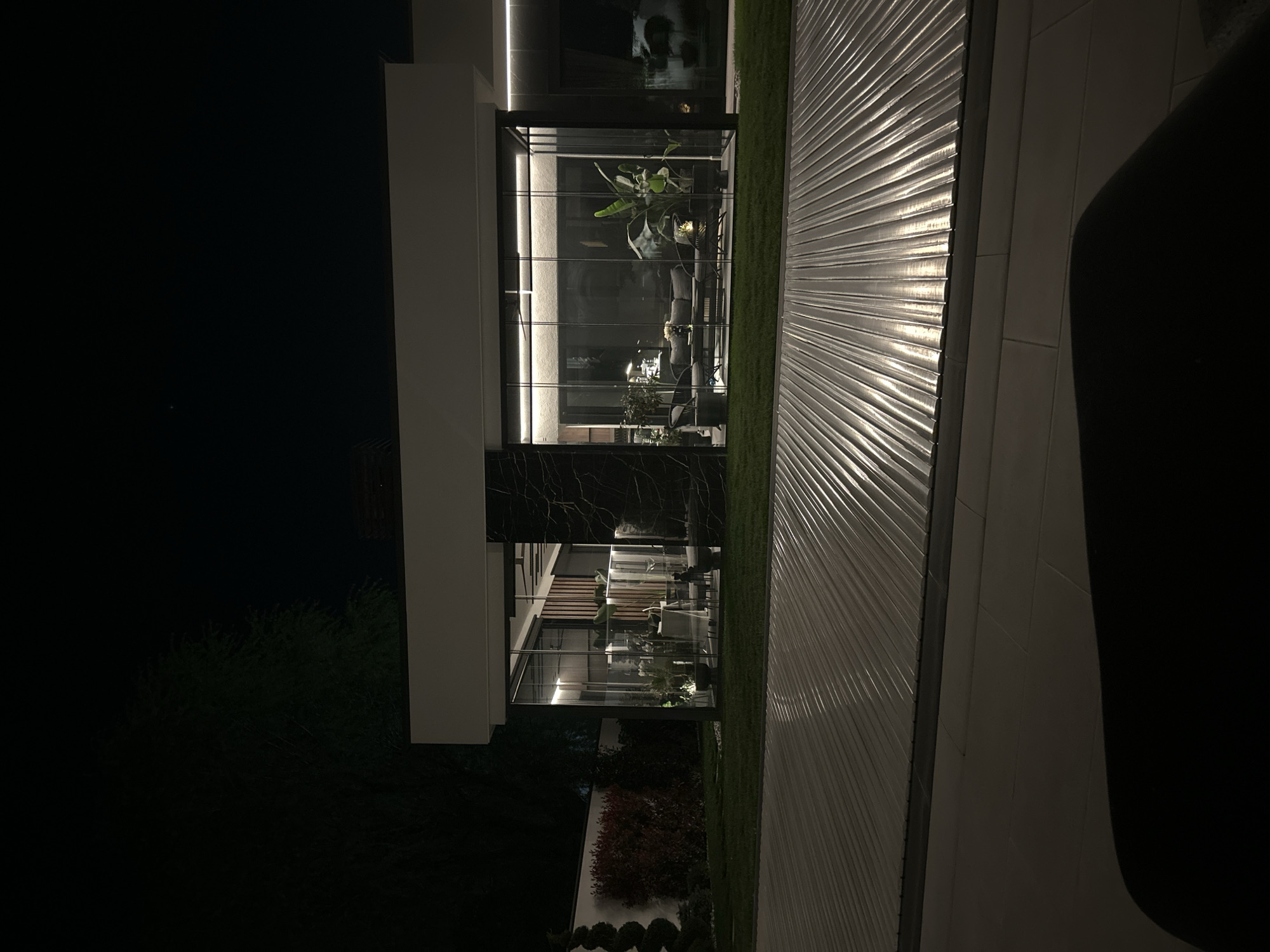 The image depicts an architectural view of a modern building at night. The building features a glass facade with several floors. Inside, plants and furniture are visible. A light-colored, textured wall runs parallel to the building. The environment includes greenery and some dark areas, likely representing trees or landscape features. The image is oriented vertically, and the location is Plovdiv, Bulgaria.