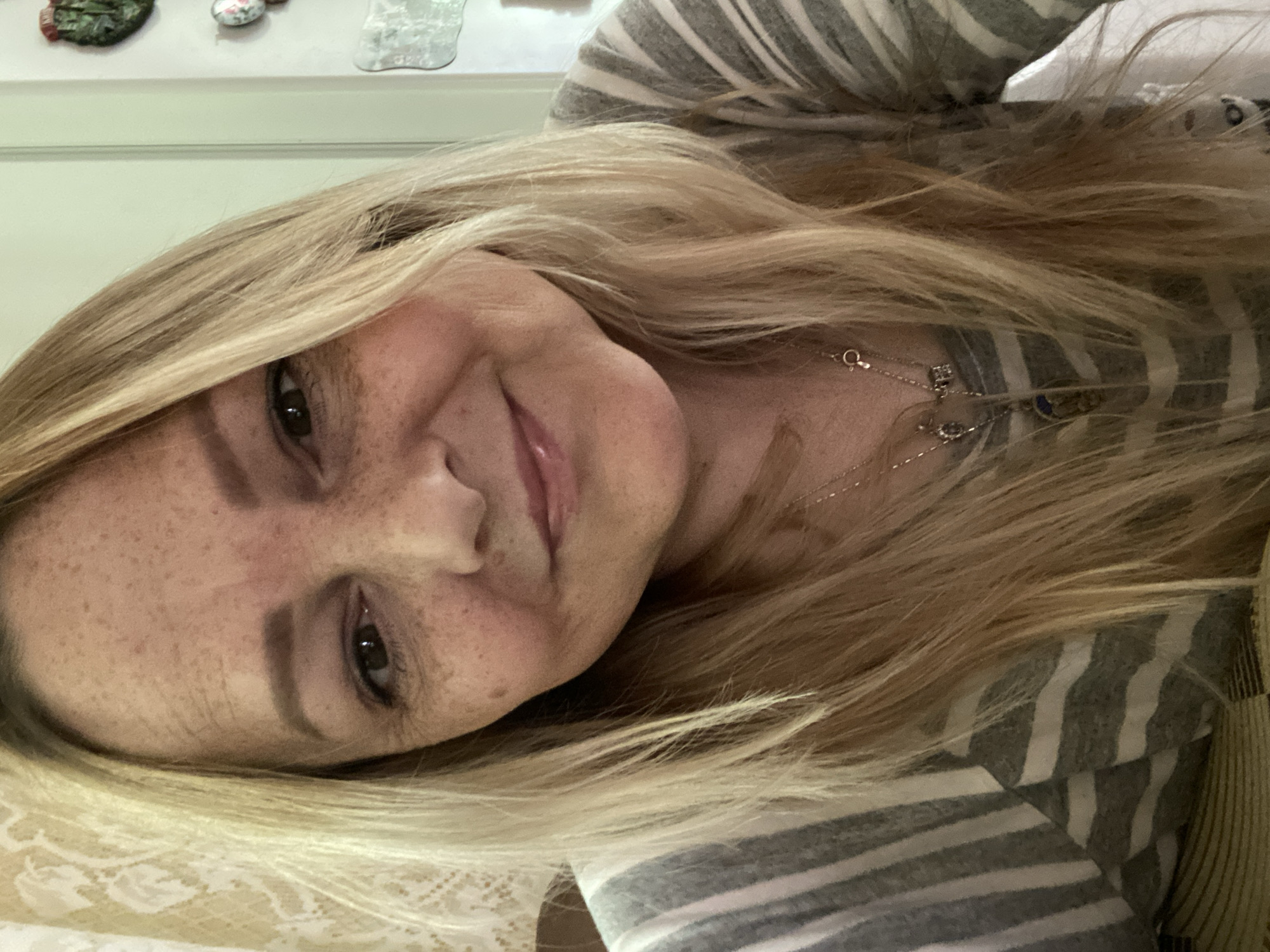 A close-up, selfie-style photograph shows a Caucasian woman with blonde hair and freckles. She is wearing a gray and white striped shirt and a necklace. The background consists of a lace curtain and a white wall with decorative items. The lighting suggests an indoor setting. The location is not specified within the city of Luh, Ukraine.