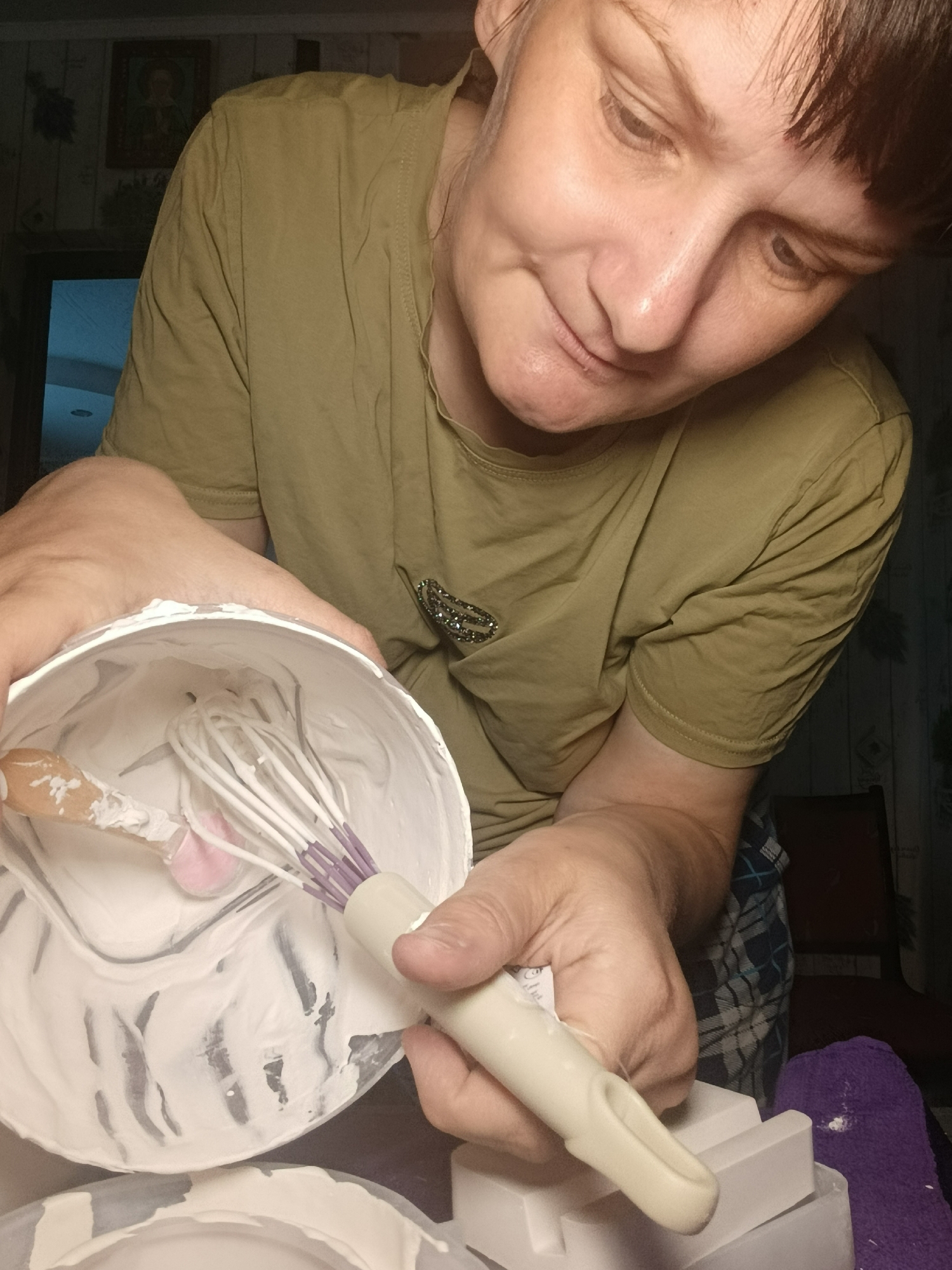 A middle-aged woman with short dark hair and an olive green t-shirt looks down into a white bucket filled with white cream. She holds the bucket with her left hand and a white and purple whisk with her right. A wooden spoon is also in the bucket. The woman is indoors, with a plaid shirt visible and a purple towel. The exact location in the city of Фоки, Russia cannot be determined from the image.