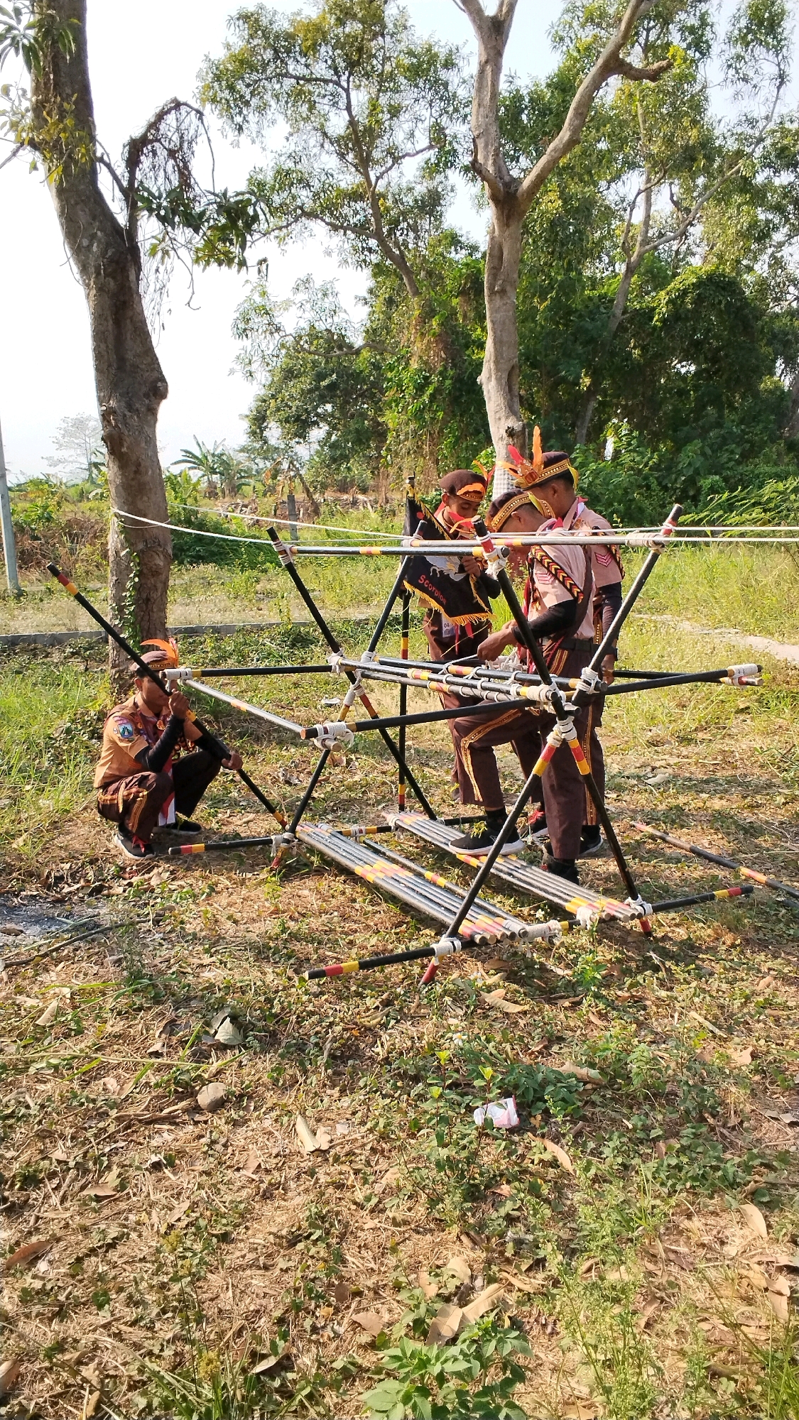 In Mojokerto, Indonesia, three individuals in brown scout uniforms and traditional headwear are assembling a bamboo structure outdoors. The structure is a complex arrangement of black bamboo poles connected by white bindings, forming a geometric framework. One scout is crouched on the ground, working on the lower section of the structure, while the other two stand, engaged in its construction. The ground is covered with dry grass and fallen leaves, suggesting a natural or park-like setting. Tall trees with green foliage form a backdrop, and the sky above is bright and clear. The overall scene depicts a scout activity, likely involving engineering or survival skills, taking place in a sunny, outdoor environment.