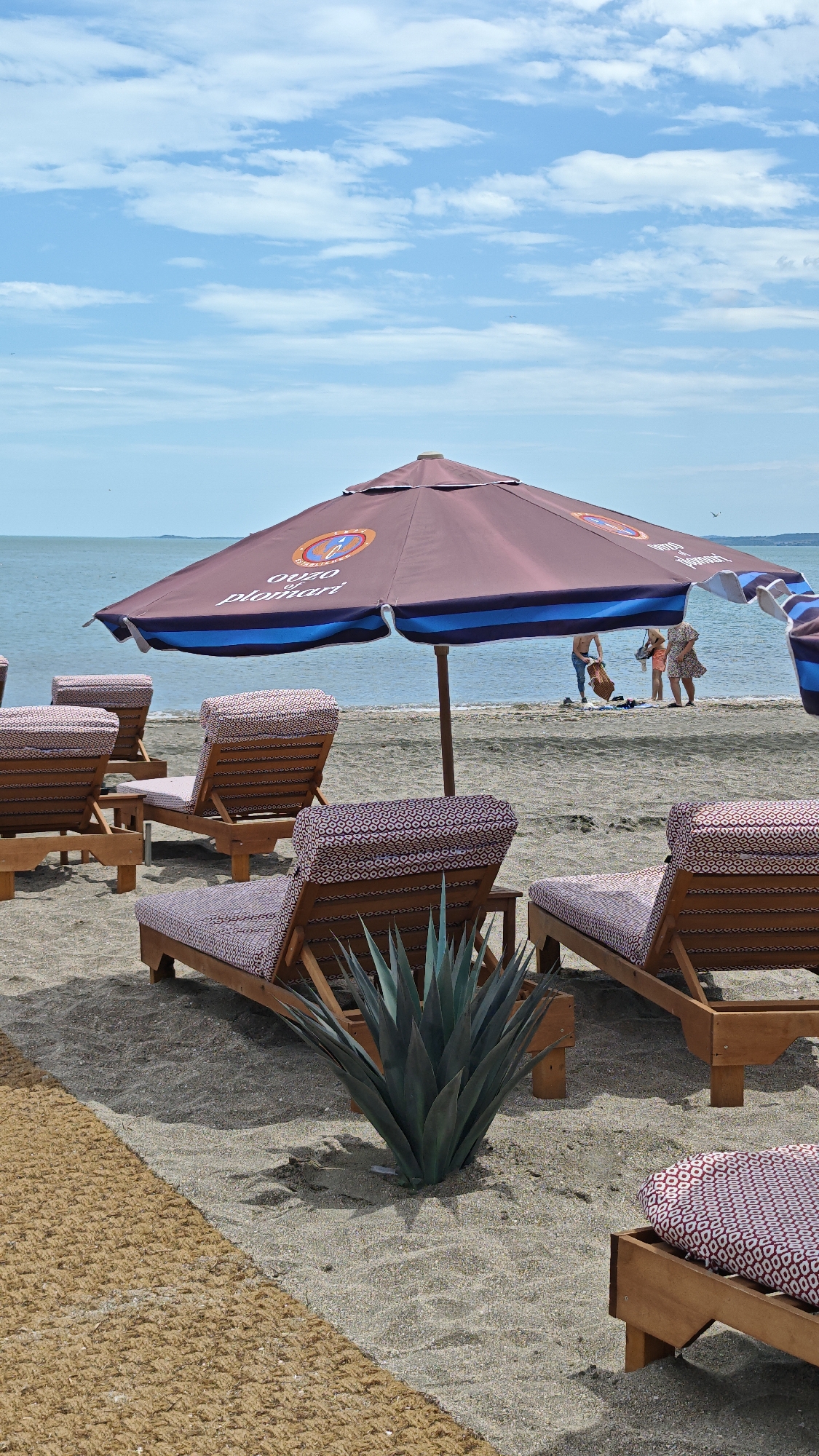 The image depicts a beach scene with several wooden lounge chairs with patterned cushions arranged under a large beach umbrella advertising "OUZO plomari". The umbrella has a brown canopy with a blue stripe. In the background, several people are standing near the water's edge. In the foreground, between the lounge chairs, is a large agave plant. To the left of the plant, there is a brown mat on the sand. The sky is blue with scattered clouds, and the sea is calm. The location is likely the city of Burgas, Bulgaria.