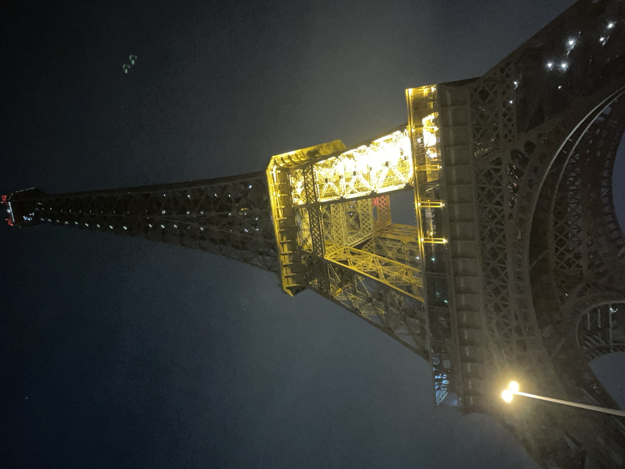 The Eiffel Tower in Paris, France is illuminated at night. The tower's metallic structure is visible against a dark sky, with warm yellow lights highlighting its intricate design and upper sections. The lower portion of the tower is in shadow, with a streetlamp providing a local light source to the left of the frame. There are two small, green lights visible in the sky to the right of the tower, and a single, distant blue light in the upper left quadrant of the image. The overall atmosphere is dark and slightly hazy, characteristic of a nighttime urban environment.