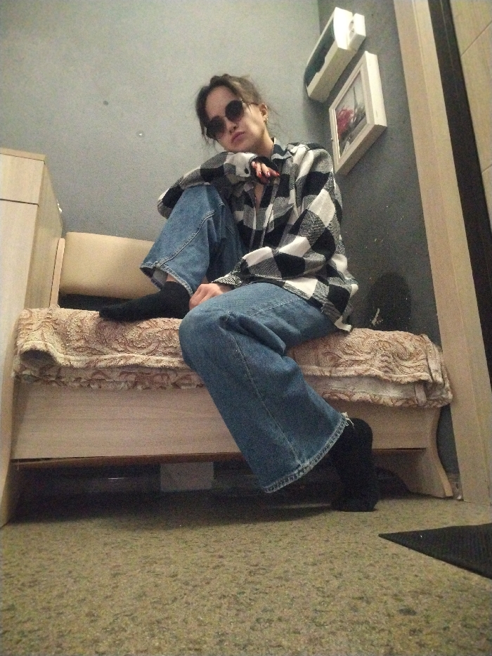 The image depicts an indoor scene within an unspecified building in Noviy, Russia. A single individual, appearing to be a young adult female, is seated on a beige upholstered bench or daybed. She is positioned towards the center of the frame, angled slightly to her left, with her head tilted. She wears an oversized black and white plaid shirt, blue denim jeans, and black socks. Dark, round sunglasses obscure her eyes, and her dark hair is pulled back. Her left hand, with red-painted fingernails, rests on her left thigh. The bench features a patterned fabric in beige and brown hues over a light wooden frame with carved details. To the left of the bench, a portion of a light-toned wooden furniture piece, possibly a cabinet, is visible. The background wall is dark gray. Mounted on this wall are a white rectangular object, potentially an electrical panel, positioned above a smaller white-framed picture depicting a scene with red accents. A dark object, consistent with a cap or small bag, rests on the floor against the wall below the framed picture. The floor is carpeted with a speckled light green and beige material. A dark rectangular mat is partially visible in the bottom right corner, adjacent to a light-colored doorframe.