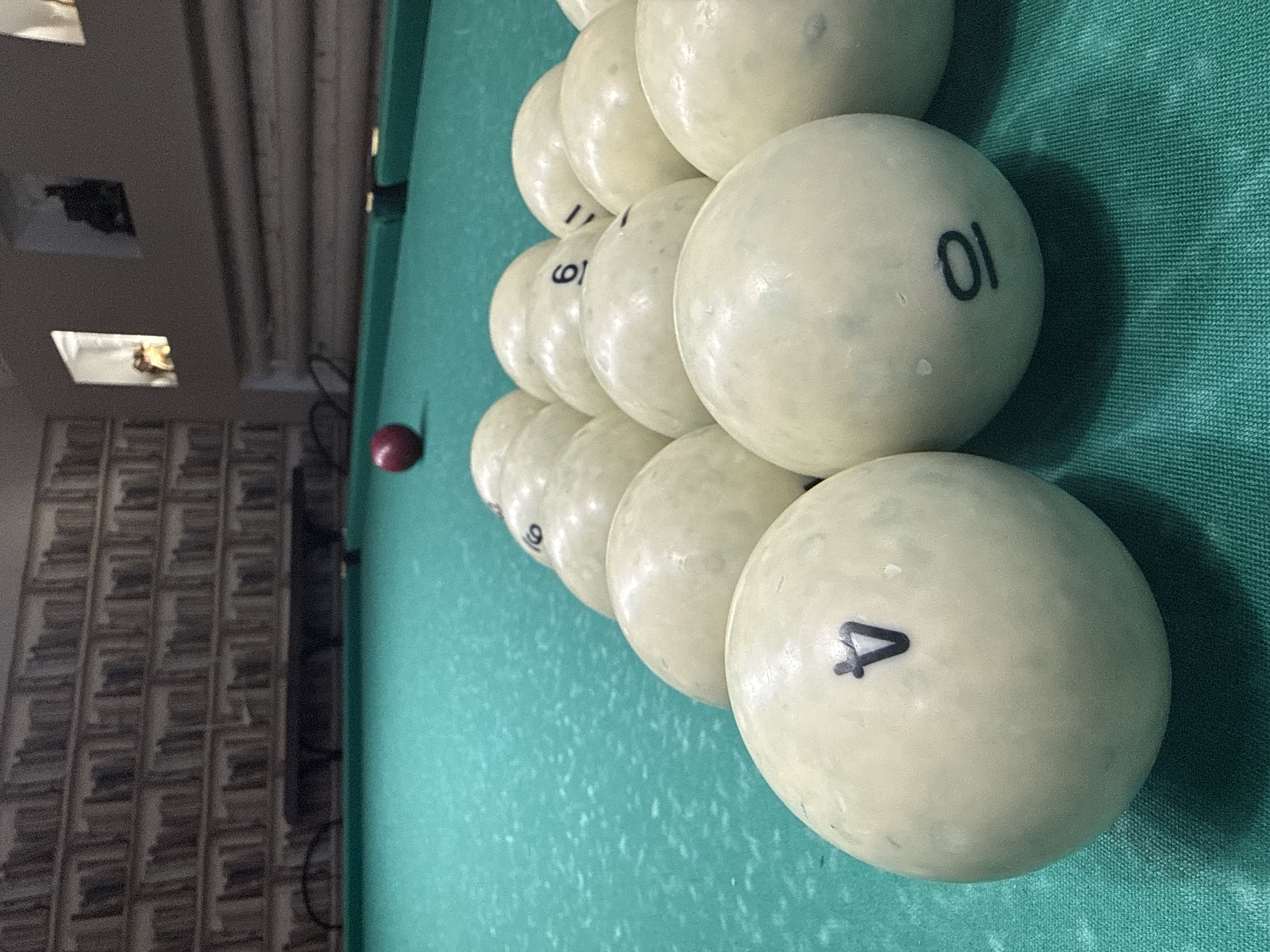 A rack of billiard balls sits on a green felt pool table. The balls are predominantly white, with some featuring black numbers. The ball closest to the viewer is white with the number "4" printed on it. Adjacent to it is a white ball with the number "01". In the background, a single red billiard ball is visible. The backdrop features a wall with a bookshelf pattern and a window with a silhouette of a cat. The location is identified as Dmitrov, Russia.