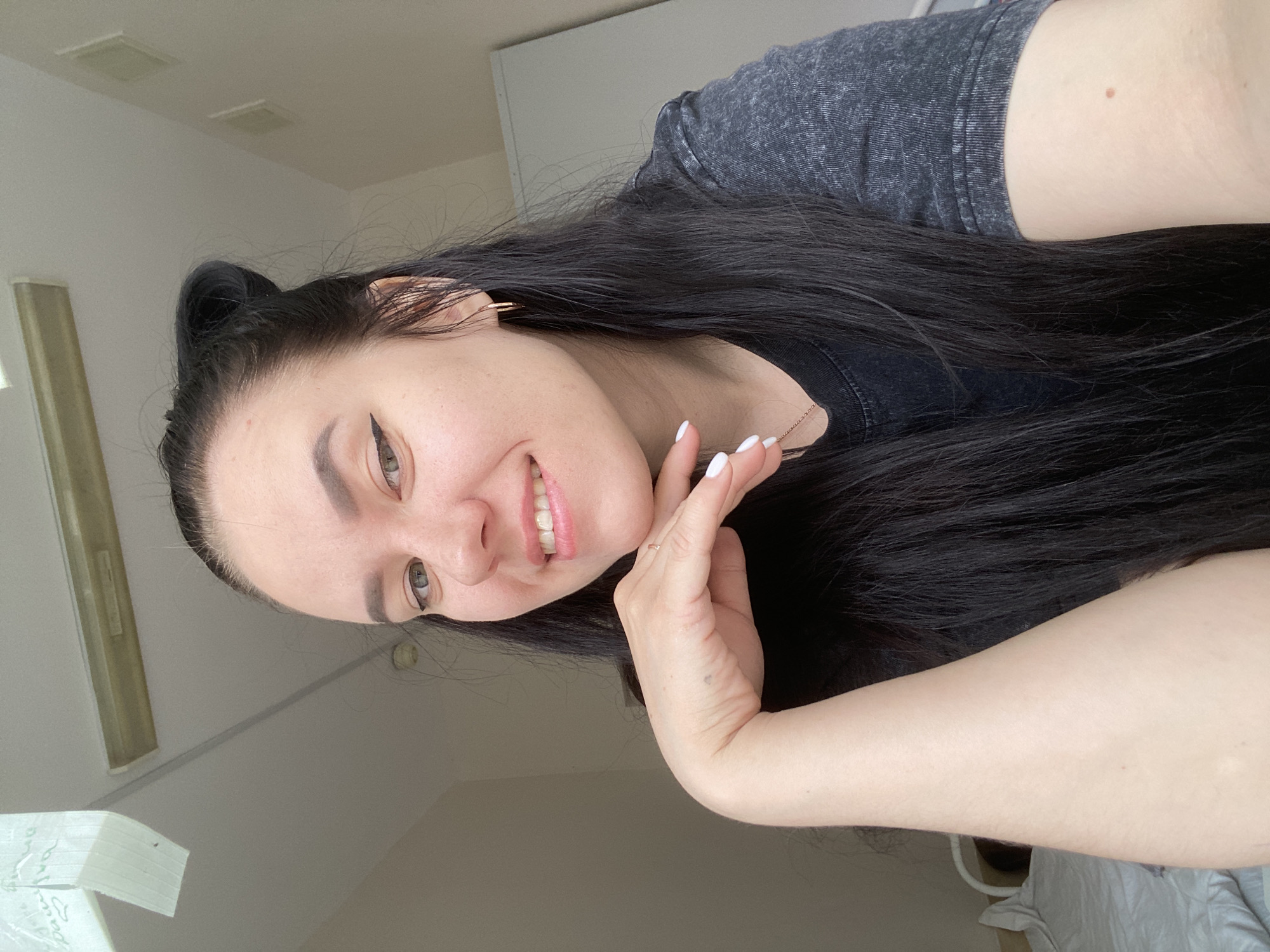 A woman with long black hair and fair skin is smiling at the camera. She is wearing a grey shirt. Her left hand is raised to her face. She has gold hoop earrings. The background is a white wall and ceiling. The location appears to be indoors, possibly a home, in Perm, Russia.