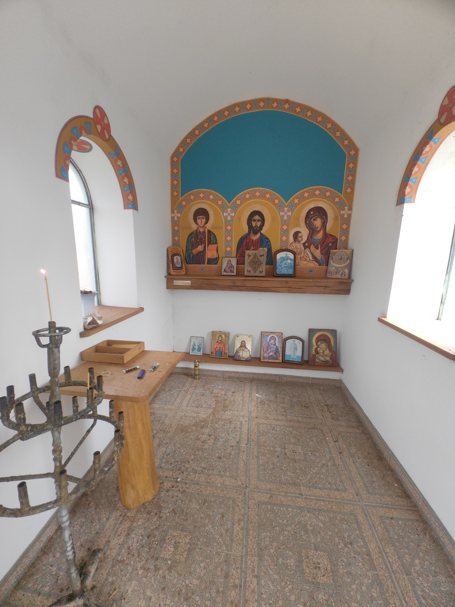The image depicts the interior of a small, likely religious, space in Gumoshtnik, Bulgaria. The room is white-walled with arched windows decorated with geometric patterns. An alcove with a teal backdrop displays three Orthodox icons. A wooden shelf beneath the icons holds smaller religious images and objects. Another row of smaller icons rests on the floor near the alcove. In the foreground, there's a rustic wooden table and a tall, ornate metal candle stand, with one candle burning. There is also a wooden tray with items on the table. The floor is tiled with an intricate, mosaic-like pattern.