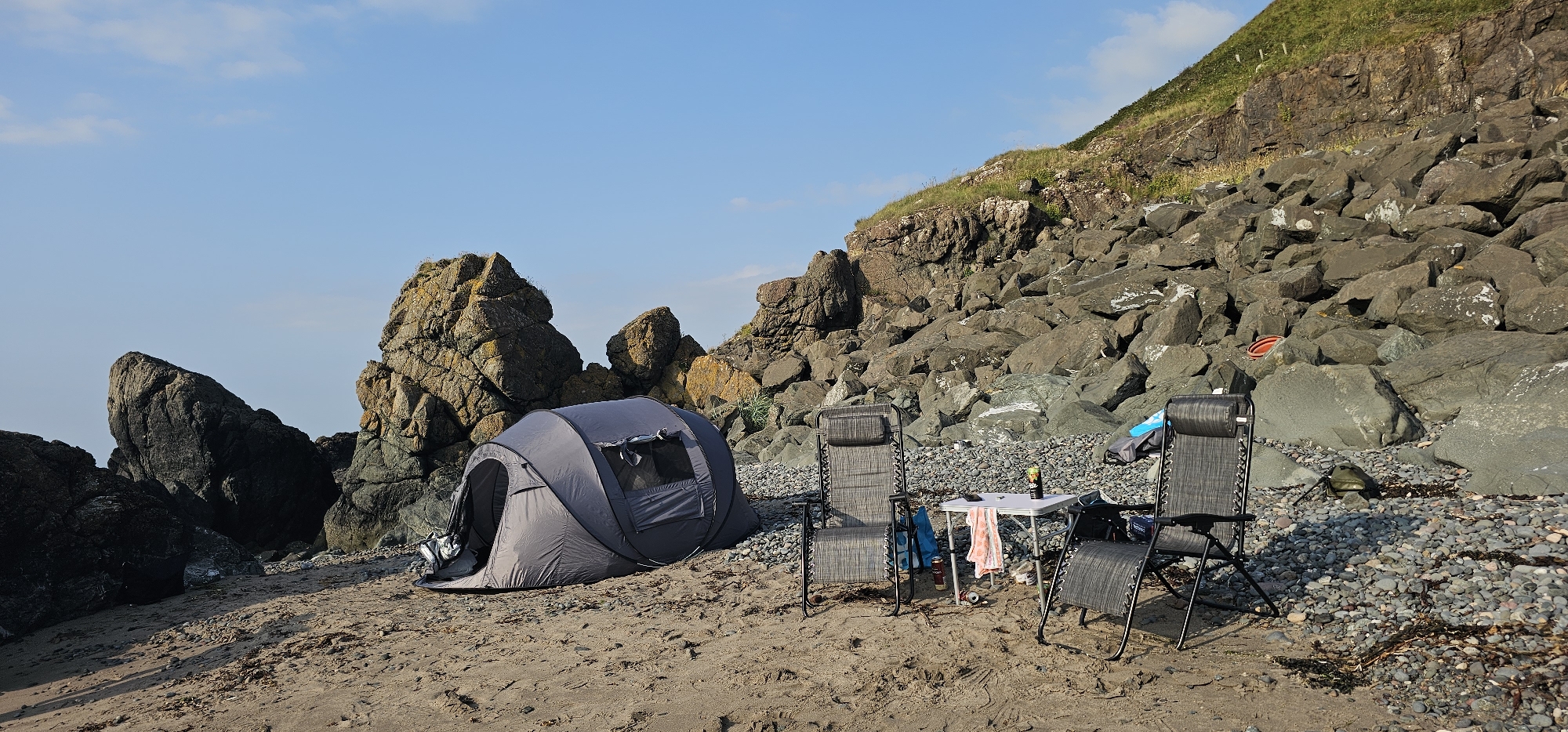 A campsite is set up on a pebble and sand beach at the base of a rocky cliff, with a blue sky overhead. In the foreground, two grey, foldable lounge chairs face a small white folding table. A grey pop-up tent is pitched to the left of the chairs, with its entrance open. On the table, there are a few bottles and a towel. The rocky cliff face is a prominent feature in the background, with sparse vegetation growing on the upper slopes. The beach is strewn with rocks and seaweed. The scene is tranquil and appears to be a location for outdoor recreation. This is likely Girvan, United Kingdom, given the coastal setting and rocky terrain common to the area.