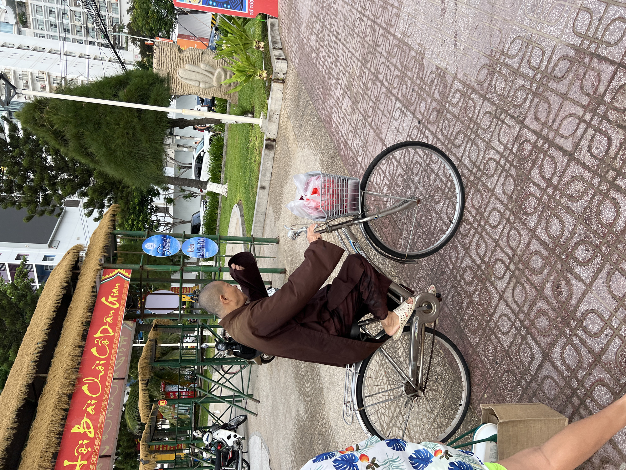 A person dressed in traditional brown Buddhist monk attire is riding a silver bicycle down a paved walkway in Nha Trang, Vietnam. The monk is facing left and appears to be looking ahead. The bicycle has a basket on the front containing red items wrapped in plastic. The walkway is paved with red and white patterned tiles. In the background, there is a building with a thatched roof and a red banner with yellow lettering. There are also trees, a lamppost, and other people visible in the distance. To the left of the monk, a person wearing a white shirt with a blue and green floral pattern is partially visible. The scene appears to be outdoors in a public area, possibly a street or park.