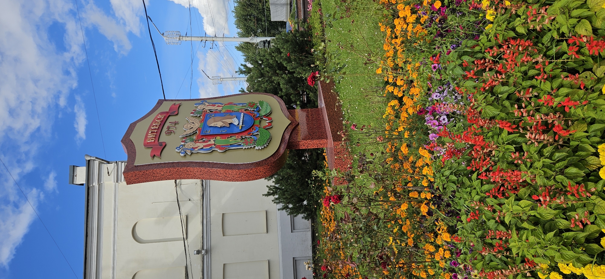 The image shows a coat of arms in the foreground, likely a city emblem, with the name "ВИТЕБСК" (Vitebsk) and the year "974" inscribed on it. The emblem is mounted on a red, granite-like pedestal. Surrounding the emblem is a vibrant flowerbed filled with a variety of colorful flowers, including red, orange, pink, and purple blooms, as well as green foliage. In the background, a light-colored building with arched windows is visible, along with some evergreen bushes and trees. Utility poles and wires are also present in the background, suggesting an urban or semi-urban setting. The sky is blue with scattered white clouds, indicating fair weather. This scene is likely located in the city of Vitebsk, Belarus, as indicated by the text on the emblem.