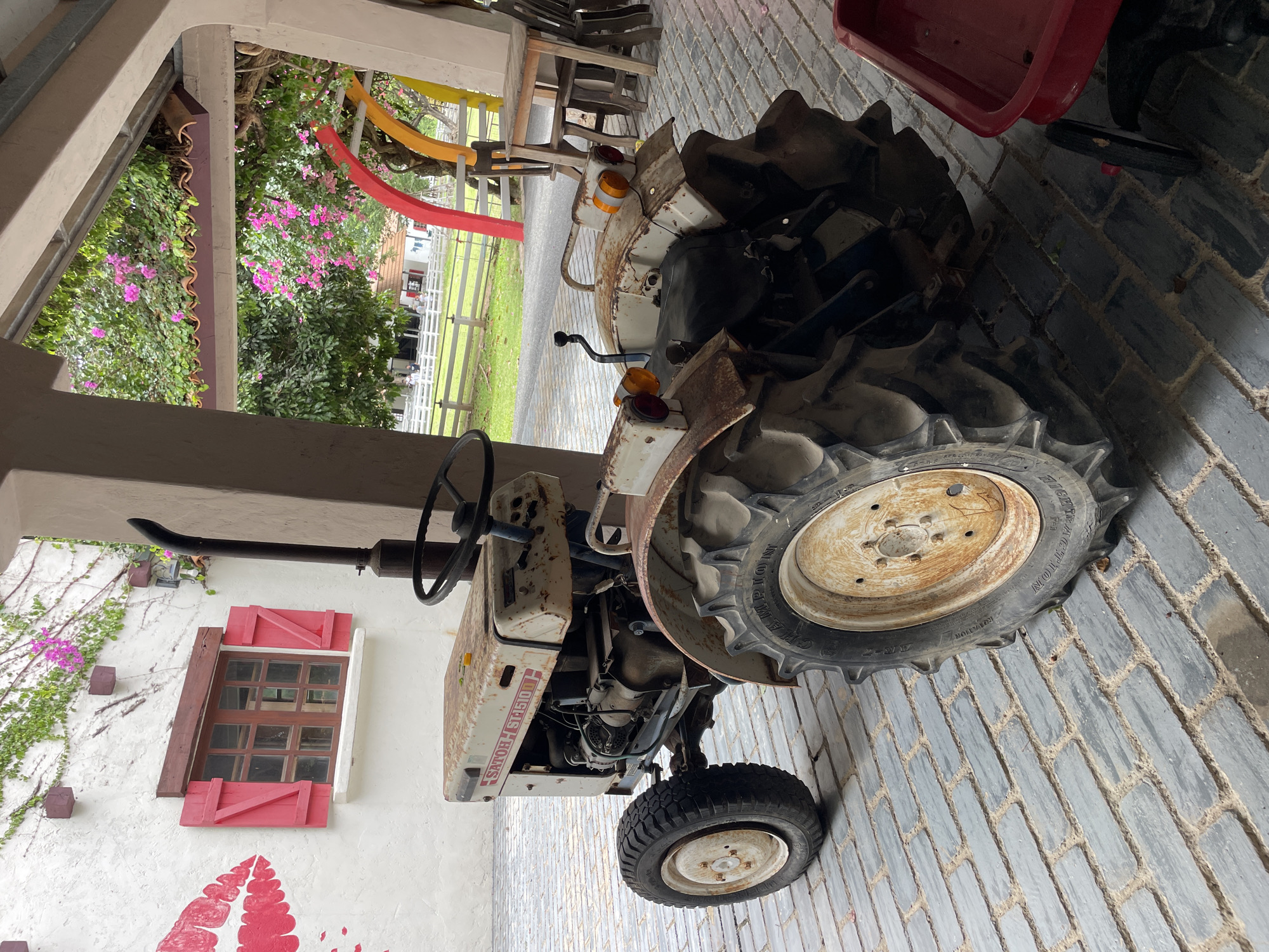 This image depicts an aged, white and red SATOR ST5101 tractor parked on a grey brick patio. The tractor is weathered and rusty, with large, rugged tires. To the left of the tractor is a white wall adorned with a large red lipstick graphic and a window with red shutters. Above the wall, green vines with pink flowers are growing. Behind the tractor, an open-air structure with a tiled roof is visible, supported by columns. Through the structure, a grassy area with a white fence and trees can be seen. The environment suggests a rural or farm-like setting, possibly in or near Na Chom Thian, Thailand. The tractor appears stationary, and no people are actively interacting with it.