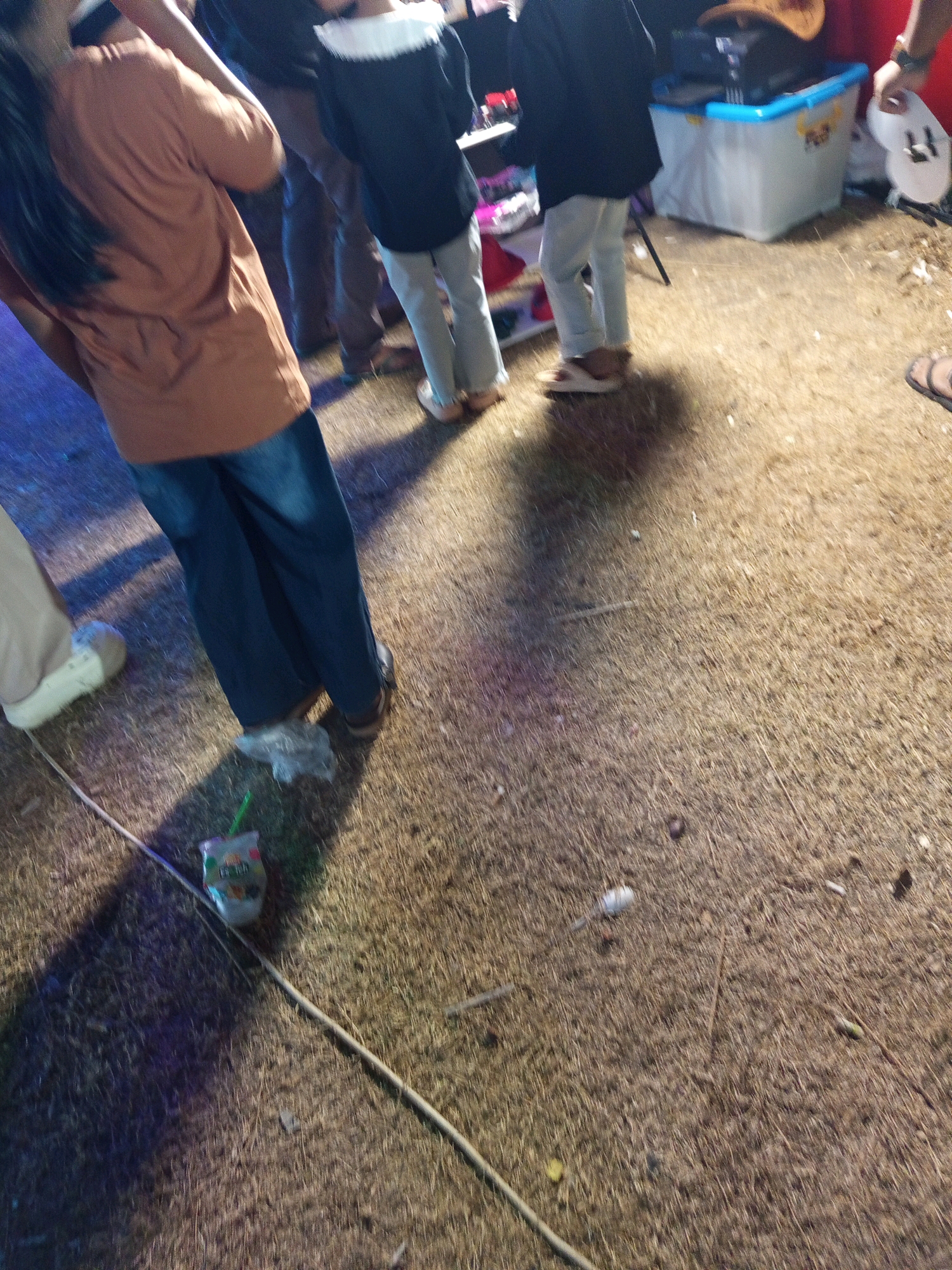 The image captures a street scene in Pulung Kencana, Indonesia, likely at dusk or during an evening event. In the foreground, a person wearing a brown shirt and blue jeans is standing, with a drink container and straw on the ground nearby. To their right, another individual is visible from the waist down, wearing light-colored pants and sandals. Further in the background, a person in a dark jacket with a white collar and light-colored pants is standing near a blue and white cooler. The ground is covered in dry grass or straw, with some scattered debris. The overall lighting suggests artificial illumination from nearby sources.
