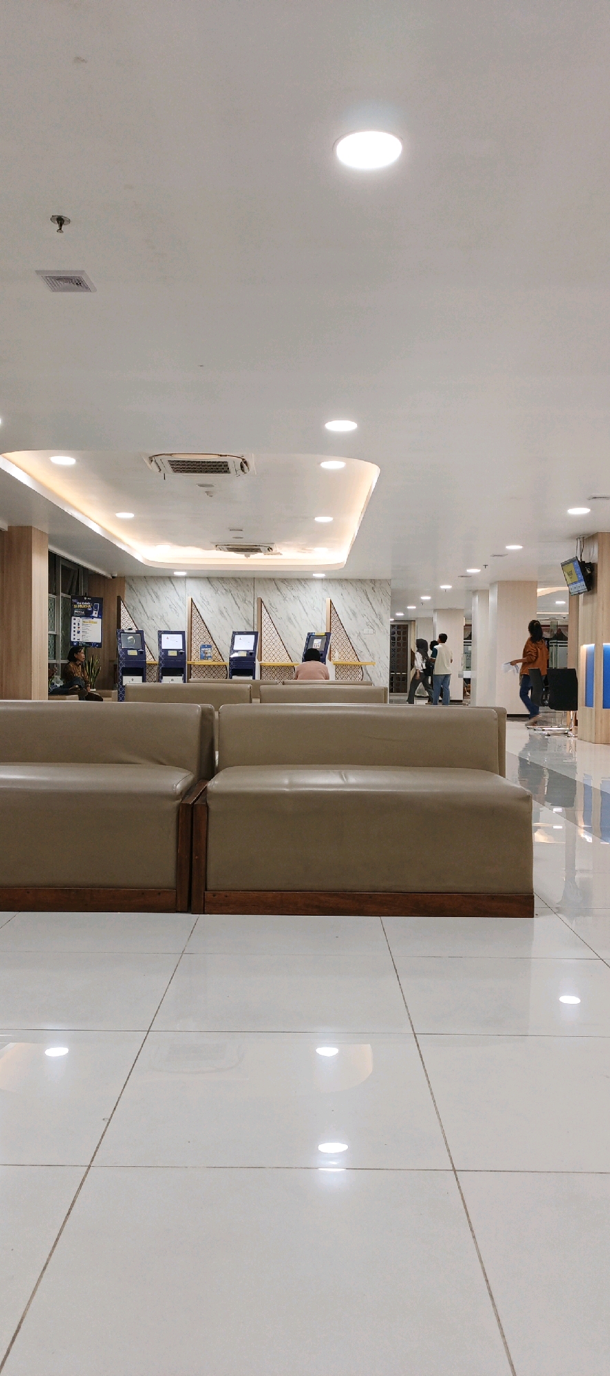 This is an interior shot of a modern waiting area, likely a bank or government office, in Trihanggo, Indonesia. The scene is well-lit with recessed ceiling lights and a decorative light fixture above a curved ceiling. In the foreground, two light brown, modular sofas with wooden frames face the viewer, their polished surfaces reflecting the bright lights.
Behind the sofas, a row of blue and silver self-service kiosks or ATMs are lined up against a marble-patterned wall. A few individuals are visible in the mid-ground, interacting with the kiosks or waiting. To the right, further down a hallway, more people are seen, including a person in an orange shirt walking away from the camera. The floor is made of large, white, polished tiles that gleam under the lights. The overall impression is one of a clean, organized, and functional public space.