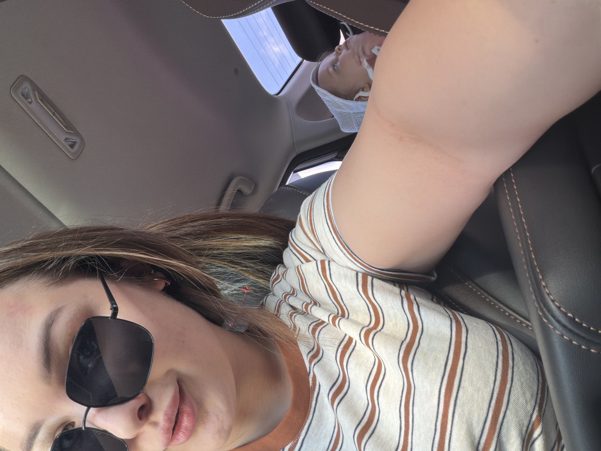 A selfie taken from a low angle inside a car. The person in the foreground is wearing sunglasses and a striped shirt. In the background, a baby is lying down, wearing a white head covering and a mask. A sunroof is visible above the baby. The car's interior is beige and black. The location is not discernible from the image.