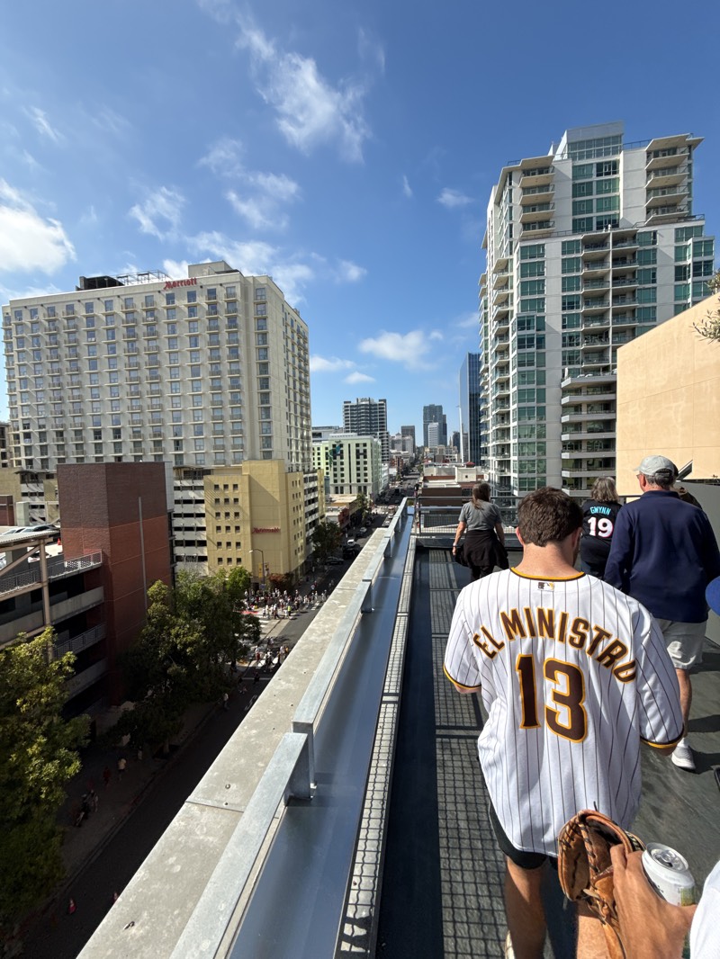 Here is a detailed description of the image:
**General Scene:**
The image captures an elevated view from what appears to be a rooftop or balcony overlooking a city street. The setting is outdoors under a blue sky with scattered clouds.
**Location (San Diego, United States):**
Based on the architecture, identifiable hotels, and crowd density, the location is most likely in downtown San Diego, California. The buildings on either side and in the distance have architectural styles consistent with the area.
**People:**
* Several individuals are visible in the foreground, all walking along a narrow, elevated pathway.
* One man in the foreground is wearing a white and brown striped San Diego Padres baseball jersey with the number "13" and the name "EL MINISTRO" on the back.
* A woman with a "Gwyn 19" jersey is walking ahead of them.
* Further ahead, there is a small crowd of people walking along the street below.
**Objects:**
* Prominent buildings include:
* A Marriott hotel is on the left side of the frame.
* Modern high-rise apartment buildings are visible on the right side.
* Other objects include:
* A railing in the foreground.
* Streetlights and utility poles are visible on the street below.
* A building on the right side.
* A baseball glove and a can.
**Actions/Interactions:**
* The primary action is people walking along the elevated walkway.
* The man in the baseball jersey is holding a baseball glove.
* The crowd of people below suggests some activity or gathering, possibly a parade or event.
**Environment:**
* The environment is urban, with a mix of commercial and residential buildings.
* The weather appears to be sunny and clear.
* There are trees, and vegetation lining the street.
**Overall Impression:**
The image captures a moment in a bustling urban environment during a pleasant day. The presence of baseball jerseys and the crowd activity suggest a celebratory or event-oriented atmosphere.