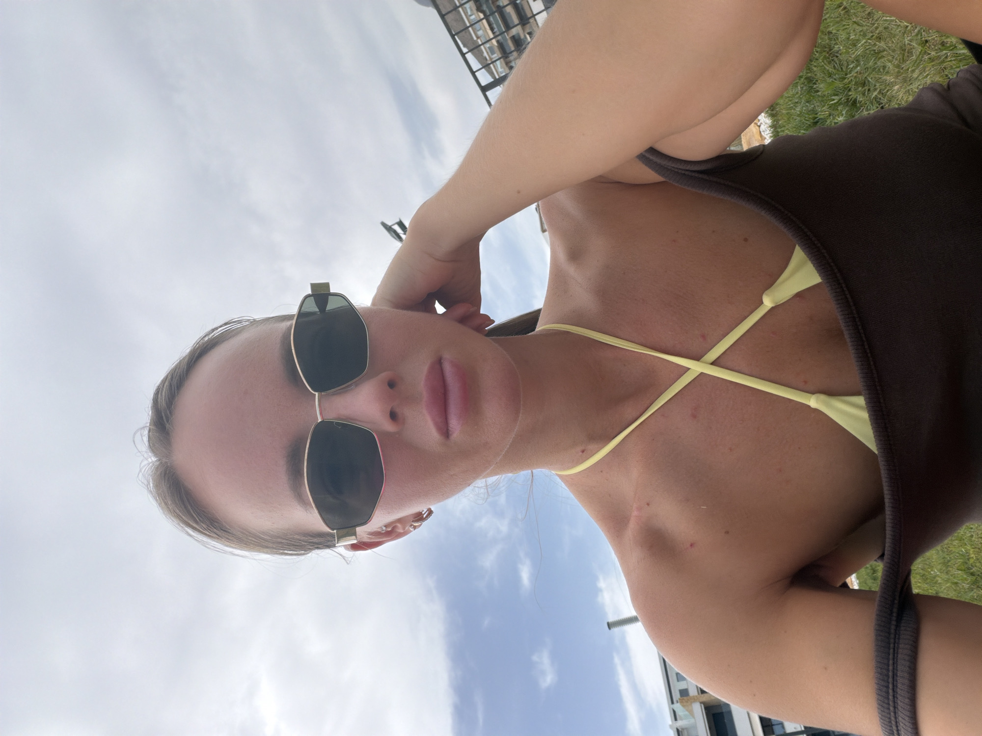 A young woman, wearing sunglasses and a bikini top with a brown tank top draped off her shoulders, sits on grass outdoors. She is looking away from the camera with her left hand resting on her head. In the background, a building is visible, suggesting an urban or resort setting. The sky is partly cloudy.