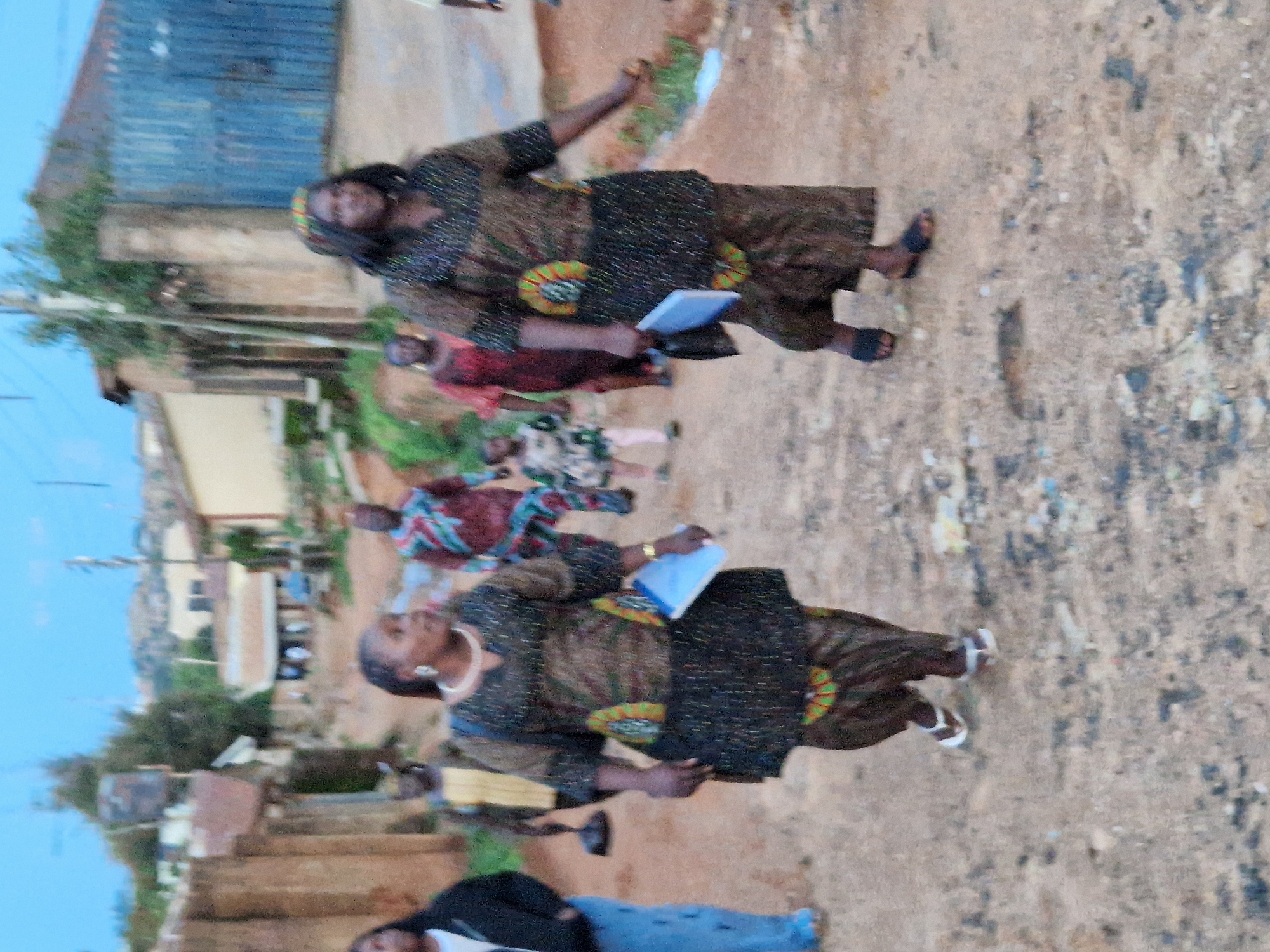 An outdoor, ground-level, slightly blurred, medium shot captures a group of people walking along a dirt road in what appears to be a residential area. The sky is overcast and a pale blue. Buildings with corrugated metal roofs and unpainted walls line both sides of the road, with some appearing dilapidated. The ground is a dusty, reddish-brown dirt with scattered debris.
In the foreground, two women are walking towards the camera. The woman on the left wears a dark, patterned outfit with a white undershirt. She has dark hair pulled back and is wearing earrings and a necklace. She is holding a light blue document. The woman on the right is dressed in a similar dark, patterned outfit, also with a patterned headscarf. She has her right arm extended slightly as she walks.
Behind them, a child in a light-colored, patterned dress is walking. Further back and to the left, a man in a yellow shirt is visible, and to the right, a person in a colorful, patterned shirt is walking away from the camera. Another person in a patterned top and patterned pants is visible further in the background. The overall impression is of a community gathering or procession in a rural or semi-urban setting.