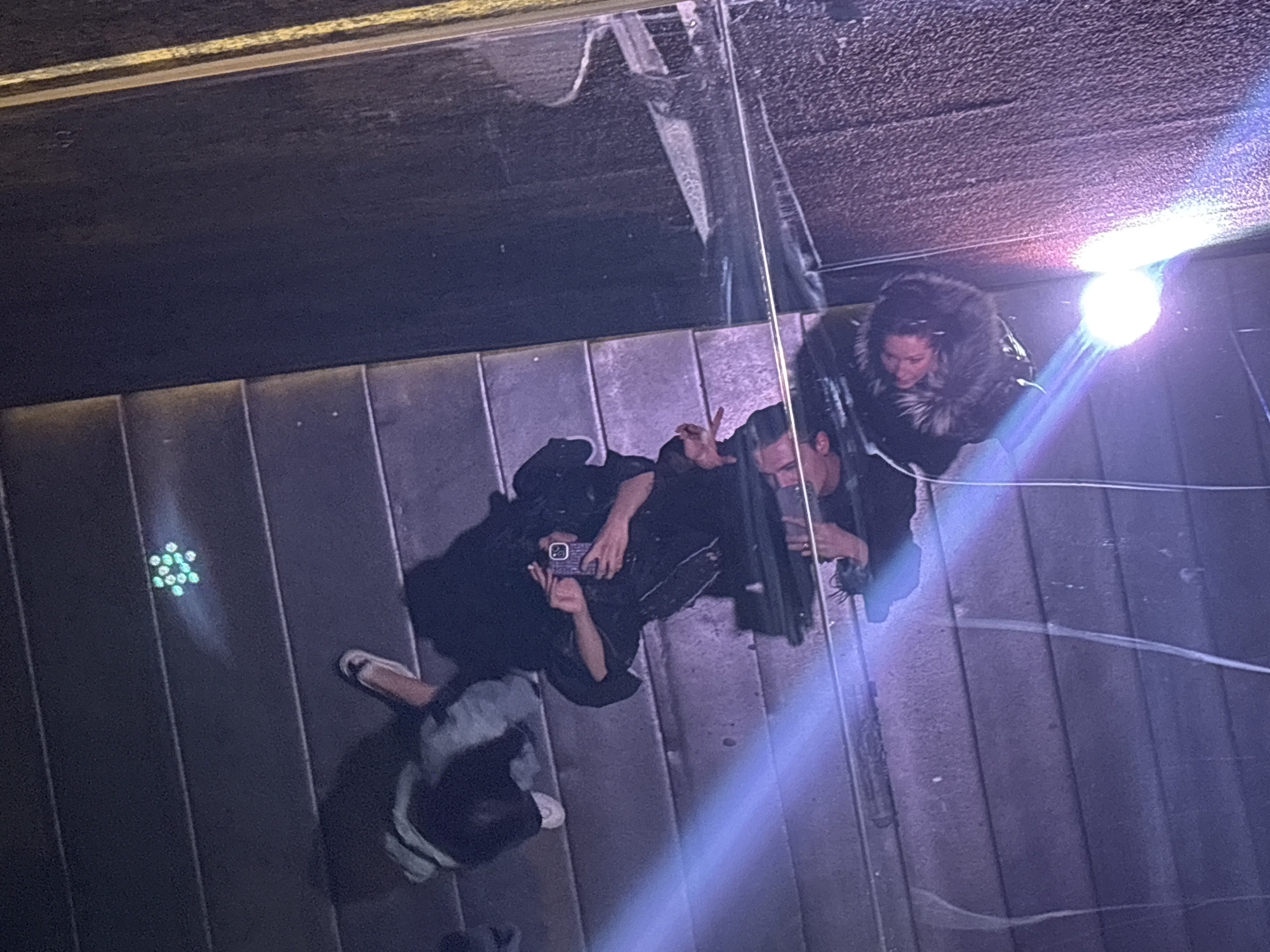 The image captures a high-angle view, likely a reflection on a polished surface or through a glass ceiling, showing three individuals in what appears to be a modern indoor or covered outdoor setting in Sofia, Bulgaria. The perspective is distorted, presenting the subjects as if viewed from above, upside down.
On the top right, a woman with dark hair wears a dark, fur-lined jacket or coat, looking up and smiling. Next to her, a man with dark hair is visible, making a V-sign with his fingers, suggesting a pose for a photo. A bright, intense light source on the right casts a strong lens flare diagonally across the image.
In the bottom left, another person is seen holding a smartphone, seemingly taking a picture, with the back of the phone and their hand clearly visible. This person is wearing light-colored sandals. The surface beneath them appears to be composed of dark, parallel planks or tiles. To their left, a small cluster of glowing green lights, arranged in a star or hexagon pattern, is visible.
The overall scene suggests a casual social gathering or tourist activity where individuals are posing for and taking photos. The dark ambient lighting combined with the bright artificial light source indicates it is likely nighttime or a dimly lit interior space. No specific text is visible in the image.
