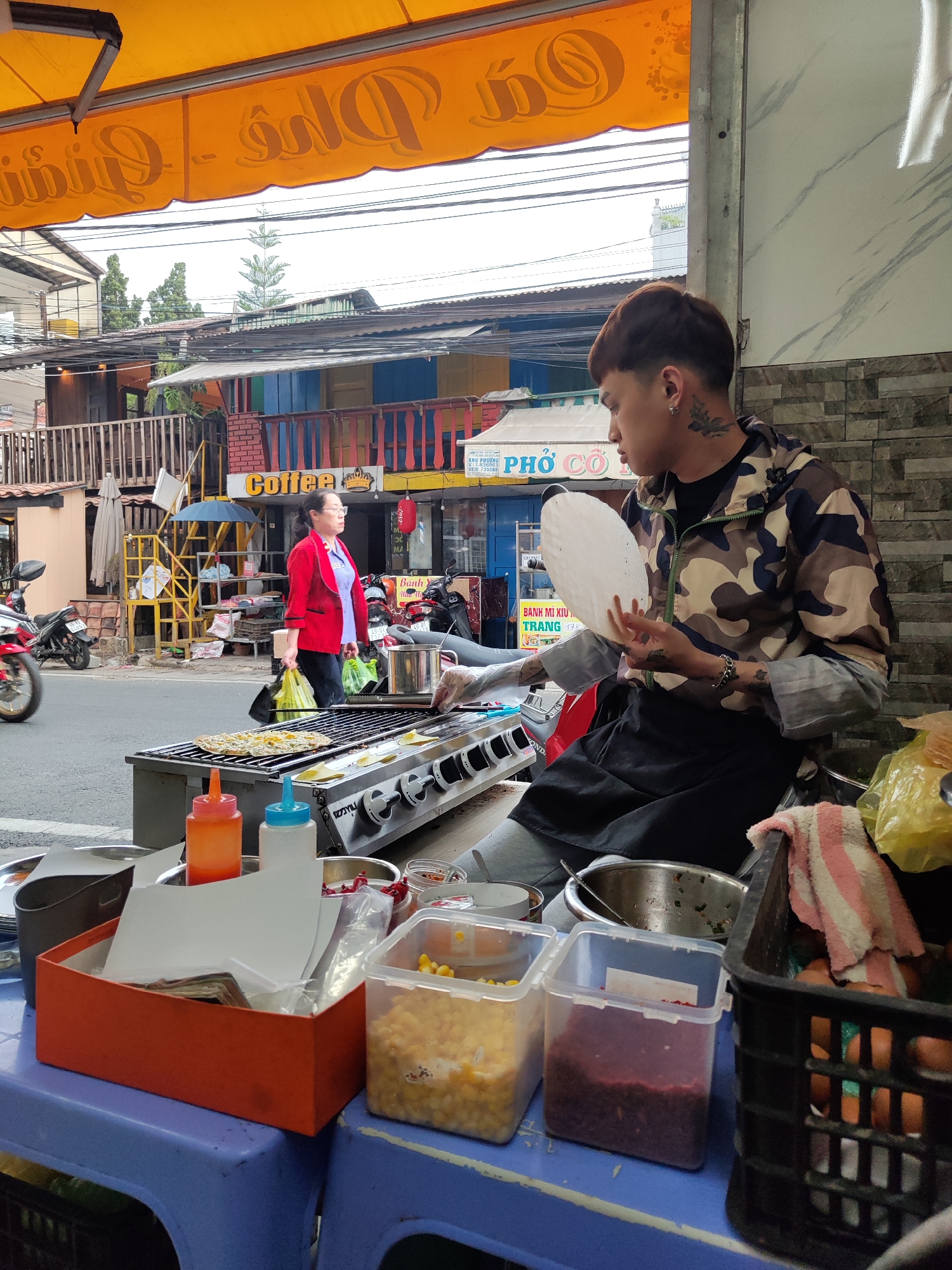 The image shows a street food vendor in Da Lat, Vietnam. A young man with tattooed neck and arms, wearing a camouflage jacket and a black apron, is seated on a motorbike. He holds a thin rice paper in his hands. In front of him is a gas grill with a thin, crepe-like food item cooking on it. Next to the grill are condiment bottles. Below the grill is a blue plastic table with several containers filled with ingredients like corn and a red condiment. An orange box is also on the table, and a black crate containing eggs is present. Behind the vendor is a wall with a faux stone pattern.
In the background, across the street, there are buildings with various signs. A woman in a red jacket is walking on the sidewalk. Motorbikes are parked along the street. The awnings of nearby businesses are visible. The overall scene suggests a bustling street food setting in Da Lat.