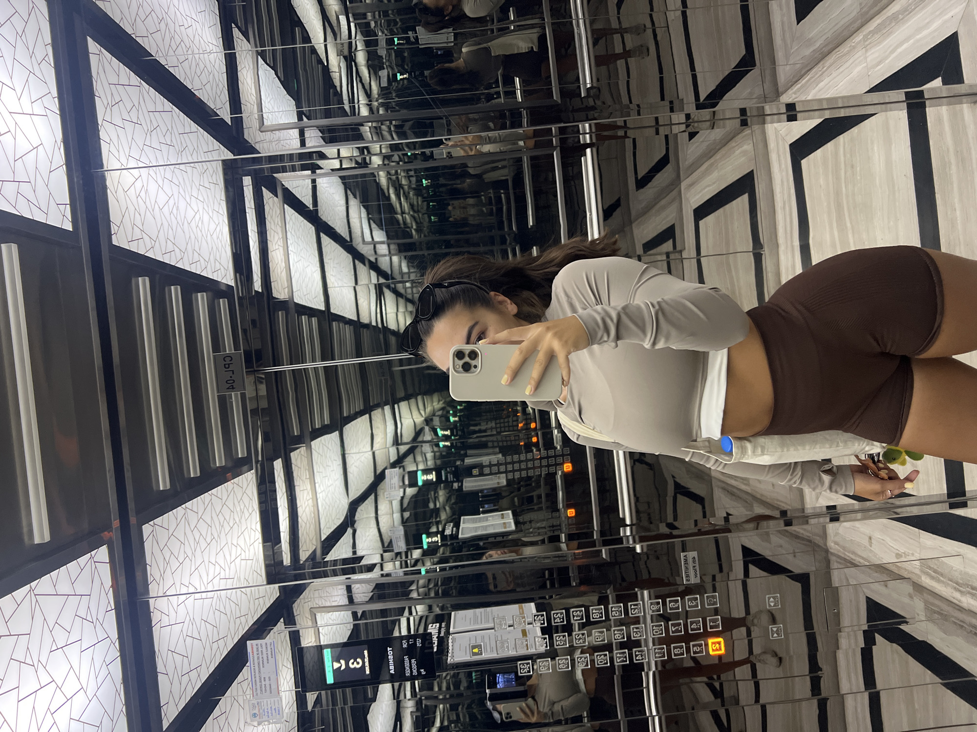 Here is a description of the image:
The image appears to be taken in an elevator with mirrored walls, creating a repeating visual effect. A person is visible in the center foreground, holding a phone and taking a selfie in the elevator mirror. The person has long hair and is wearing a light-colored long-sleeved top and brown shorts.
The elevator features a control panel with buttons and a digital display showing the floor number. The interior design of the elevator includes patterned walls and a black-and-white marble floor.
Based on the style of architecture and elevator design, the image appears to be taken in the city of Dubai, United Arab Emirates.