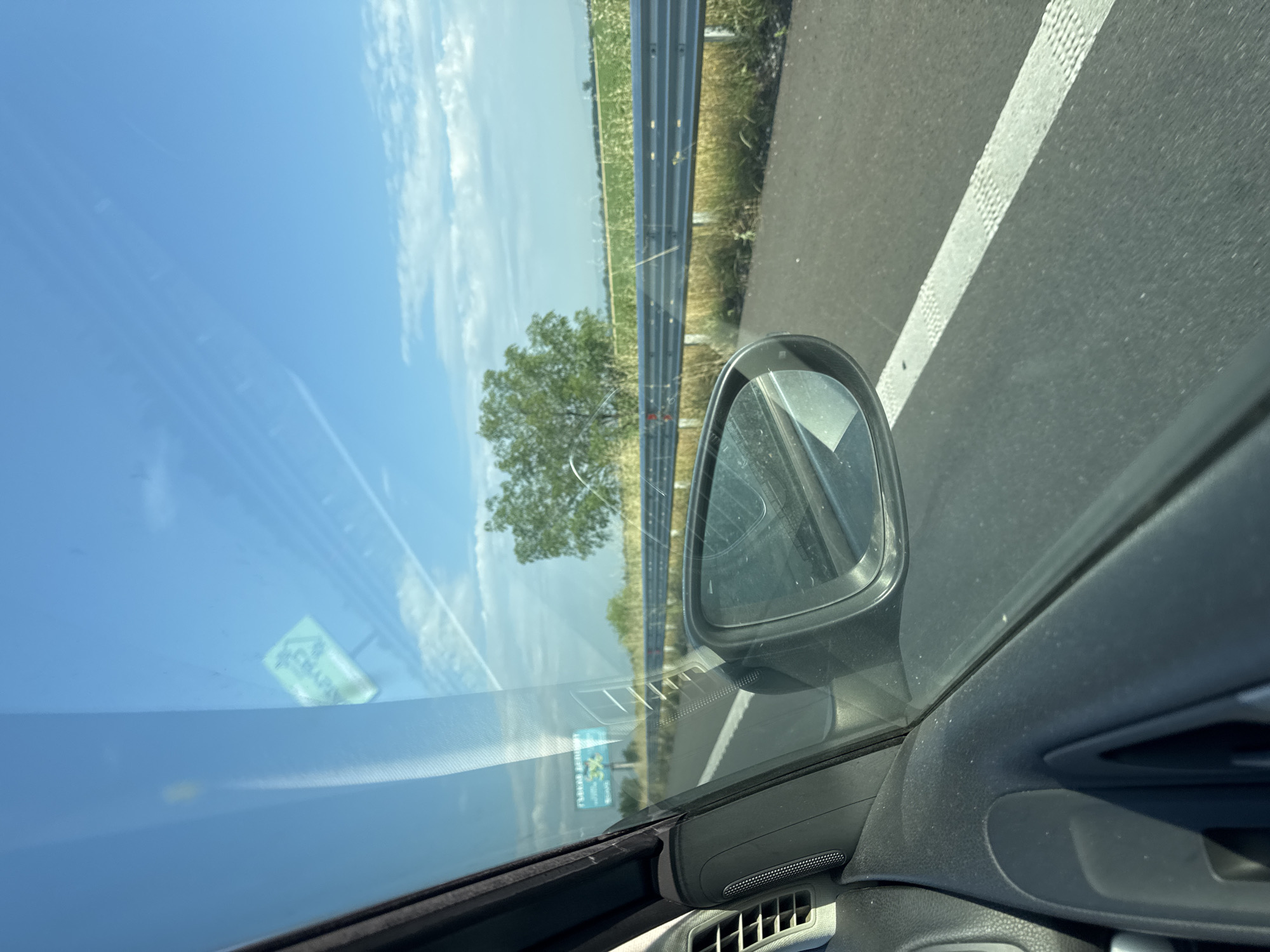 The image presents a view from the interior of a vehicle, looking outward through the passenger-side window onto a highway environment. The location is specified as Malo Konare, Bulgaria.
The sky is clear and bright blue, featuring scattered white cumulus clouds and prominent aircraft contrails in the upper right quadrant. The road visible is an asphalt highway, with a textured white dashed line delineating the right side of the lane. Adjacent to the roadway is a silver-colored metal guardrail. Beyond the guardrail, the roadside terrain consists of dry, yellowish-green grass and a solitary, verdant deciduous tree. Further into the distance, extensive agricultural fields, predominantly green with lighter patches, stretch towards the horizon. Faint outlines of distant wind turbines are discernible against the sky in the background.
The interior of the vehicle is visible in the foreground, including a segment of the black dashboard and door panel. This section features a rectangular air vent, a circular speaker grille, and a portion of the door's grab handle recess. The passenger-side exterior mirror is visible, reflecting a distorted view of the highway and parts of the car's interior. Reflections on the windshield include an inverted yellow-green sticker with partially legible Cyrillic characters, possibly "CEBAИР". A billboard with blue text, including "ГРЕДЛ" and "3%", is faintly visible through the windshield in the distance. No individuals are present in the image. The perspective and environment indicate the vehicle is in motion on the highway.