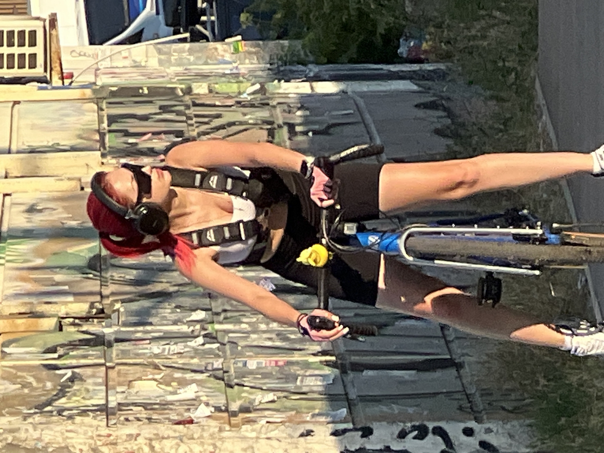 A person with bright red hair, wearing headphones, a white sports bra, black athletic shorts, and white sneakers, is riding a bicycle. They are wearing black fingerless gloves and a black harness. A yellow artificial flower is attached to the bicycle's frame. The person is leaning back slightly, with their head tilted up and eyes closed, suggesting a relaxed or enjoying moment.
The scene appears to be outdoors, possibly on a street or path. In the background, there are structures that look like they are covered in posters or graffiti, creating a colorful and textured backdrop. The sunlight suggests it is daytime, and the shadows indicate a bright, sunny day. The overall impression is one of leisure and enjoying an activity on a pleasant day. The cropped images provide a closer look at the person's clothing, accessories, and the details of the bicycle and the surrounding environment. No visible text is clearly legible in the image. The location context provided is Krasnodar, Russia.