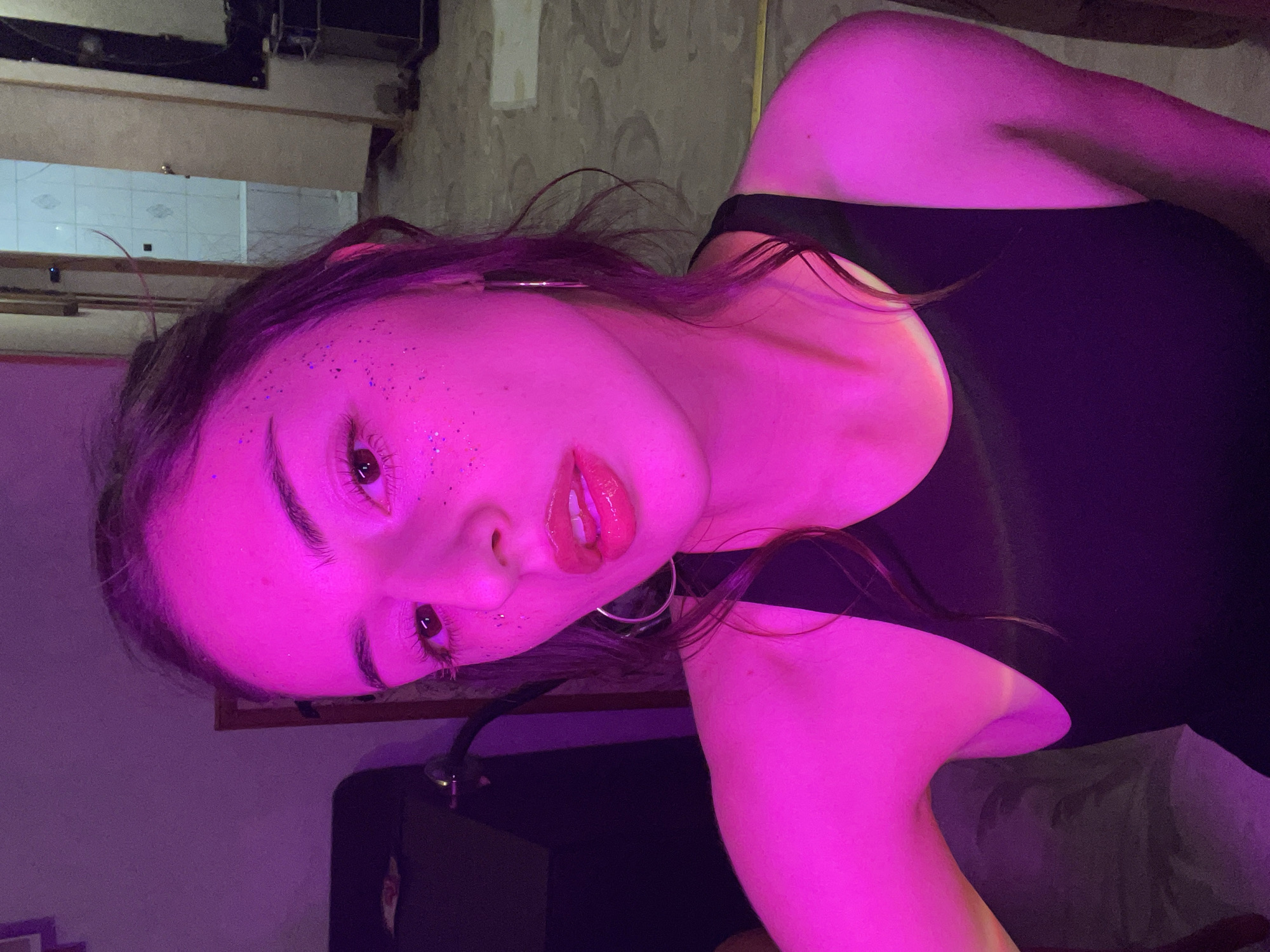 A woman is shown in a medium close-up, angled slightly downward and toward the left. The lighting is predominantly pink. The background appears to be an interior space with a combination of bare concrete walls and tiled surfaces. The scene is NOT located in Moscow, Russia.