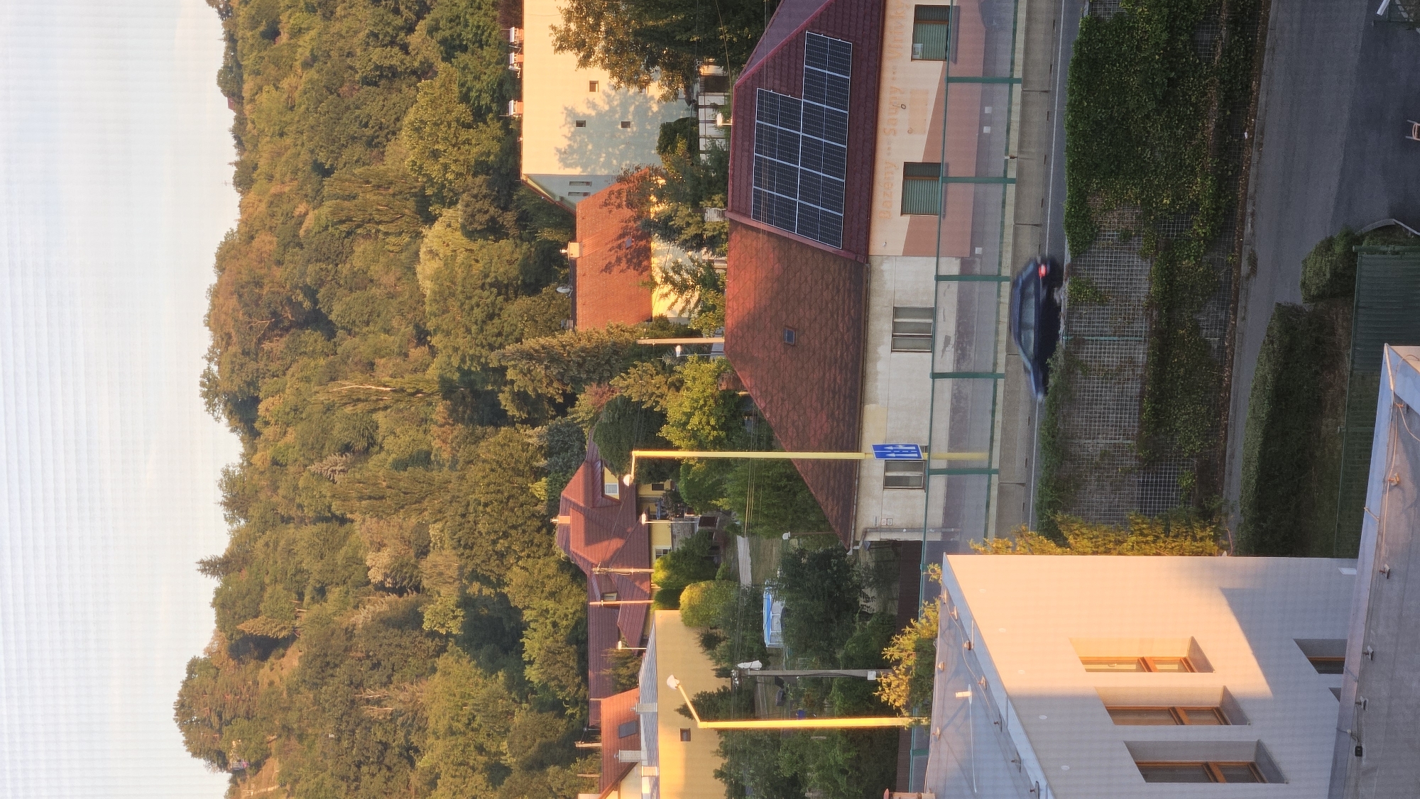 The image captures a suburban street scene in Prešov, Slovakia, bathed in the warm glow of late afternoon sunlight. The foreground features the corner of a modern, white building with large, rectangular windows reflecting the golden light. Beyond this, a street is visible, with a dark blue car moving from left to right. The car is partially obscured by a tall, green hedge lining the road, which is backed by a metal fence.
Further into the scene, a row of houses with red-tiled roofs is visible. One house in particular stands out with a prominent array of solar panels on its roof. These houses are nestled amidst lush green trees, and a gentle, tree-covered hill rises in the background, indicating the presence of natural landscape within the city. Streetlights with yellow poles are positioned along the road. In the distance, a larger, cream-colored building with multiple windows is partially visible amongst the trees. The overall impression is one of a peaceful residential area with a blend of modern and traditional architecture, surrounded by abundant greenery.