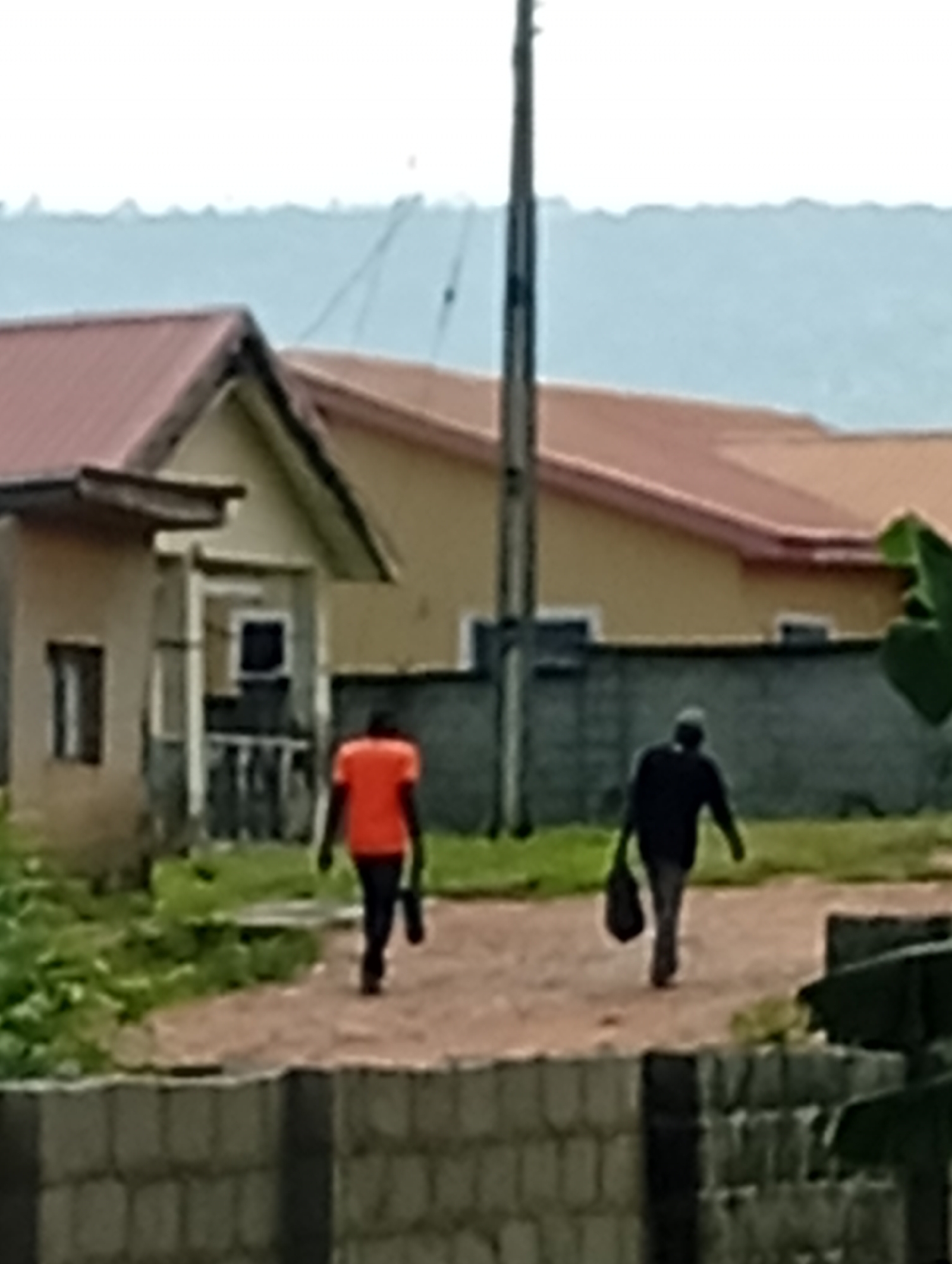 Two individuals walk away from the camera on a dirt path in front of a building with a red-tiled roof. The person on the left wears an orange shirt and dark pants, carrying a dark bag in their left hand. The person on the right wears a dark shirt and pants and carries a dark bag in their left hand. A light-colored fence is visible in the foreground. The background shows a pale sky and a distant, hazy landscape.
