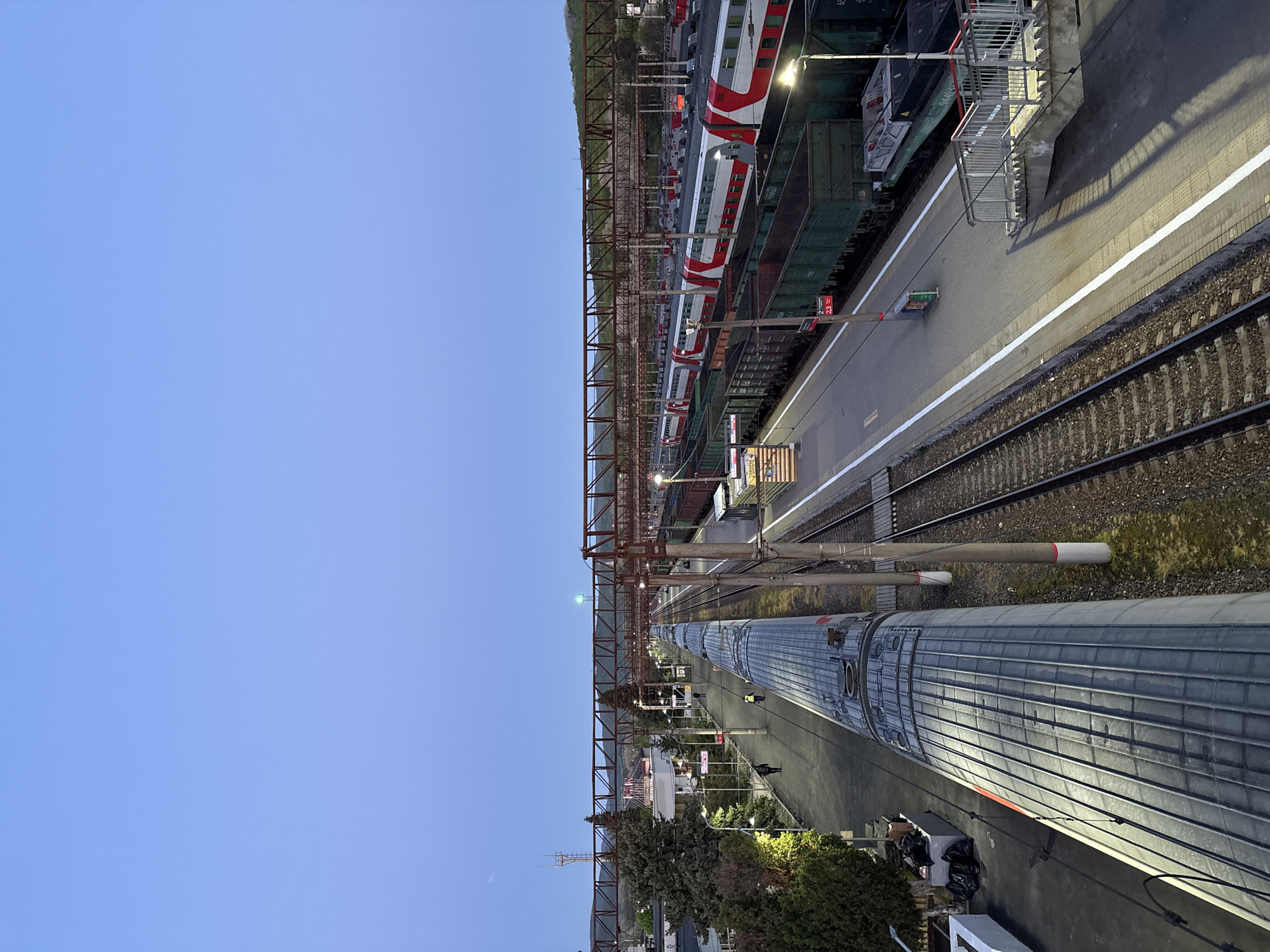 An elevated view captures a railway station or yard at twilight or night, illuminated by artificial lights under a clear, deep blue sky.
The scene features multiple parallel railway tracks. In the mid-ground on the right, a modern passenger train with red, white, and grey livery (resembling a Russian Railways "Lastochka" unit) is parked alongside a platform. The number "23" is visible on its side. Behind this passenger train, several dark green freight wagons are visible on an adjacent track, one faintly marked "9144". In the foreground on the left, a long, grey train with a ridged roof and a subtle red stripe is partially visible.
A prominent network of brown-red metal gantries and electrical catenary lines spans across the tracks, supporting the overhead power system. Lampposts cast warm light onto the platforms and tracks, creating reflections and shadows. One platform on the right, alongside the red and white train, has a white safety line and a metal staircase. A faint crescent moon is visible in the clear, deep blue sky towards the top-left. Green trees and bushes line the platforms and a distant hillside on the right. Some low-rise buildings are dimly visible in the far distance on the left.
The scene appears static and quiet, with no people visible. The trains are parked, suggesting they are either awaiting departure or are stationary in the yard. The time of day is late evening or night, indicated by the reliance on artificial lighting and the dark sky. The infrastructure and train types are consistent with a railway setting in Kaluzhskaya, Russia.