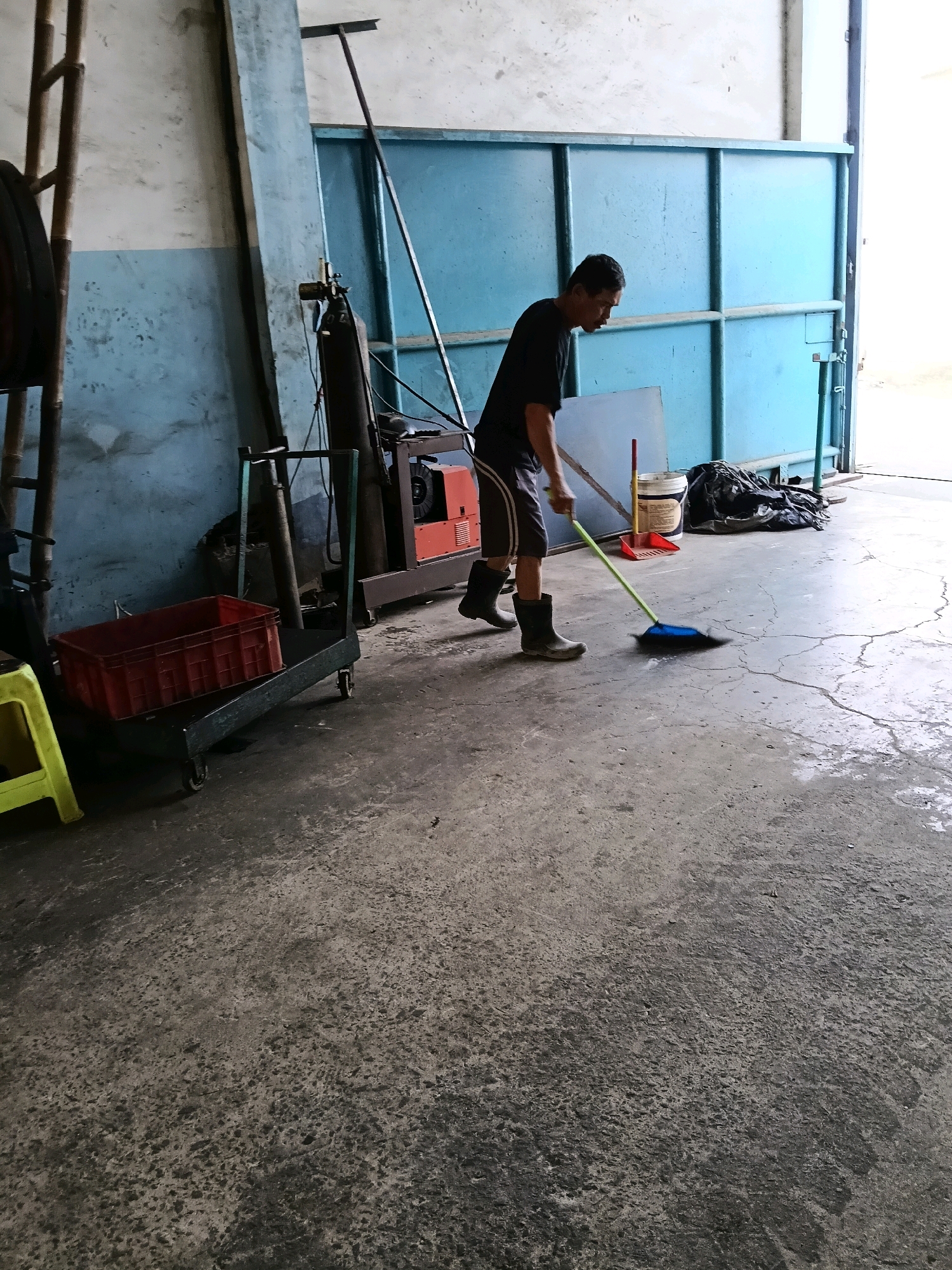 In the city of Belimbing, Indonesia, a man is sweeping the concrete floor of what appears to be a workshop or storage area. He is wearing a black t-shirt, dark shorts, and black rubber boots. He is holding a broom with a light green handle and is sweeping a dark pile of debris towards the right side of the frame. The floor is cracked and shows signs of being wet in some areas. To the left of the man, there is a red plastic crate on a green wheeled cart. Further left, a yellow plastic stool is partially visible. In the background, a large, pale blue metal door or gate is open, revealing a bright outdoor area. Behind the man, a welding machine with an orange body is visible, along with a propane tank. The walls are painted in a light blue and white pattern. A long, dark metal pole leans against the wall near the blue door.