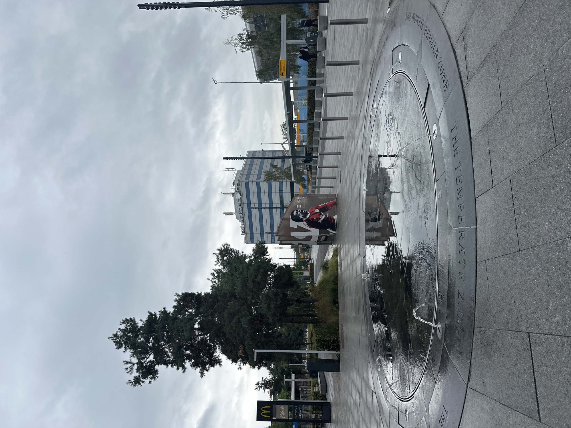 The image depicts an outdoor public space in front of a modern building in Eindhoven, Netherlands, likely Eindhoven Airport given the signage visible. In the foreground, a circular, shallow water feature with several small fountains is active, with water splashing and creating reflections on the wet, grey stone pavement. Inscribed around the rim of the water feature are the words "THE TEMPERATE ZONE" and "THE NORTH FROZEN ZONE".
To the left of the water feature, a McDonald's restaurant with a McCafe is visible, indicated by its iconic golden arches sign. Beyond that, a large evergreen tree stands prominently.
In the middle ground, a large portrait poster of a man in a red and white football jersey is displayed on a brown structure. Behind this, a modern, multi-story building with a blue and white facade bears the text "Eindhoven Airport". Further to the right, a covered shelter with a "Taxi pick up point" sign is visible, and several people are gathered or walking in this area. The sky is overcast and grey, suggesting a cool, perhaps damp, day. The overall environment appears to be a well-maintained public plaza or transportation hub.