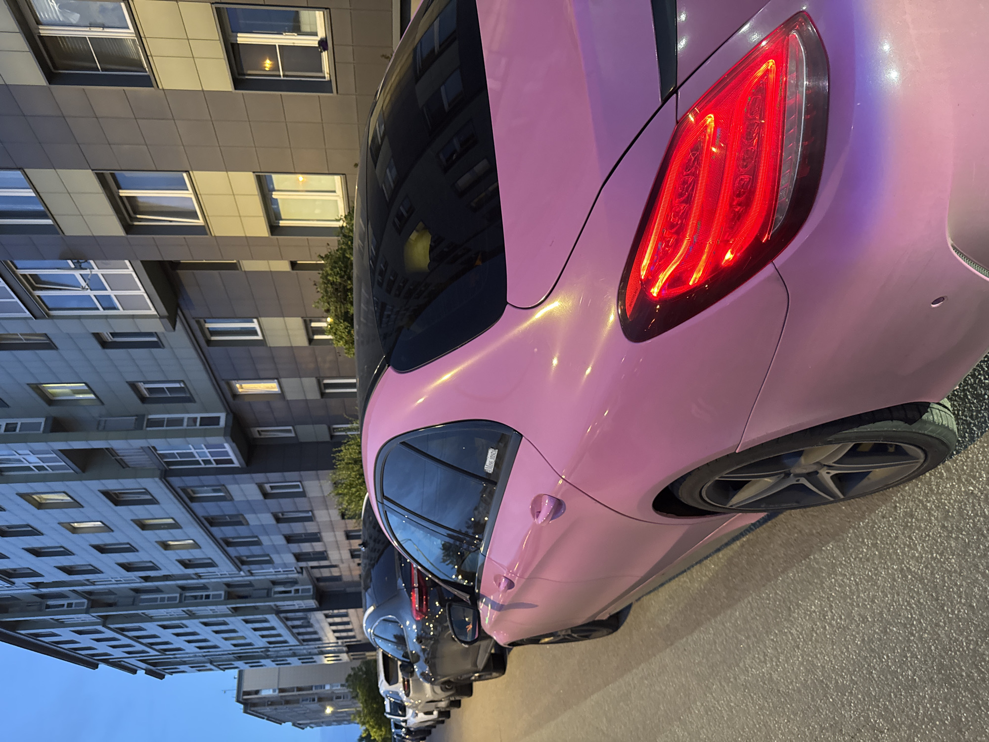 The image depicts a row of parked cars on a street in Shushary, Russia, with a modern apartment building in the background. The foreground is dominated by the rear of a pink car, which has a black roof and tinted windows. Its taillight is illuminated. To its left, a black SUV and several other vehicles are parked in a line along the curb. The apartment building is multi-story with a facade featuring a combination of grey and lighter colored panels, and numerous windows, some of which are lit. The sky is dark, suggesting it is either early morning or evening. The scene is static, with no visible human activity.