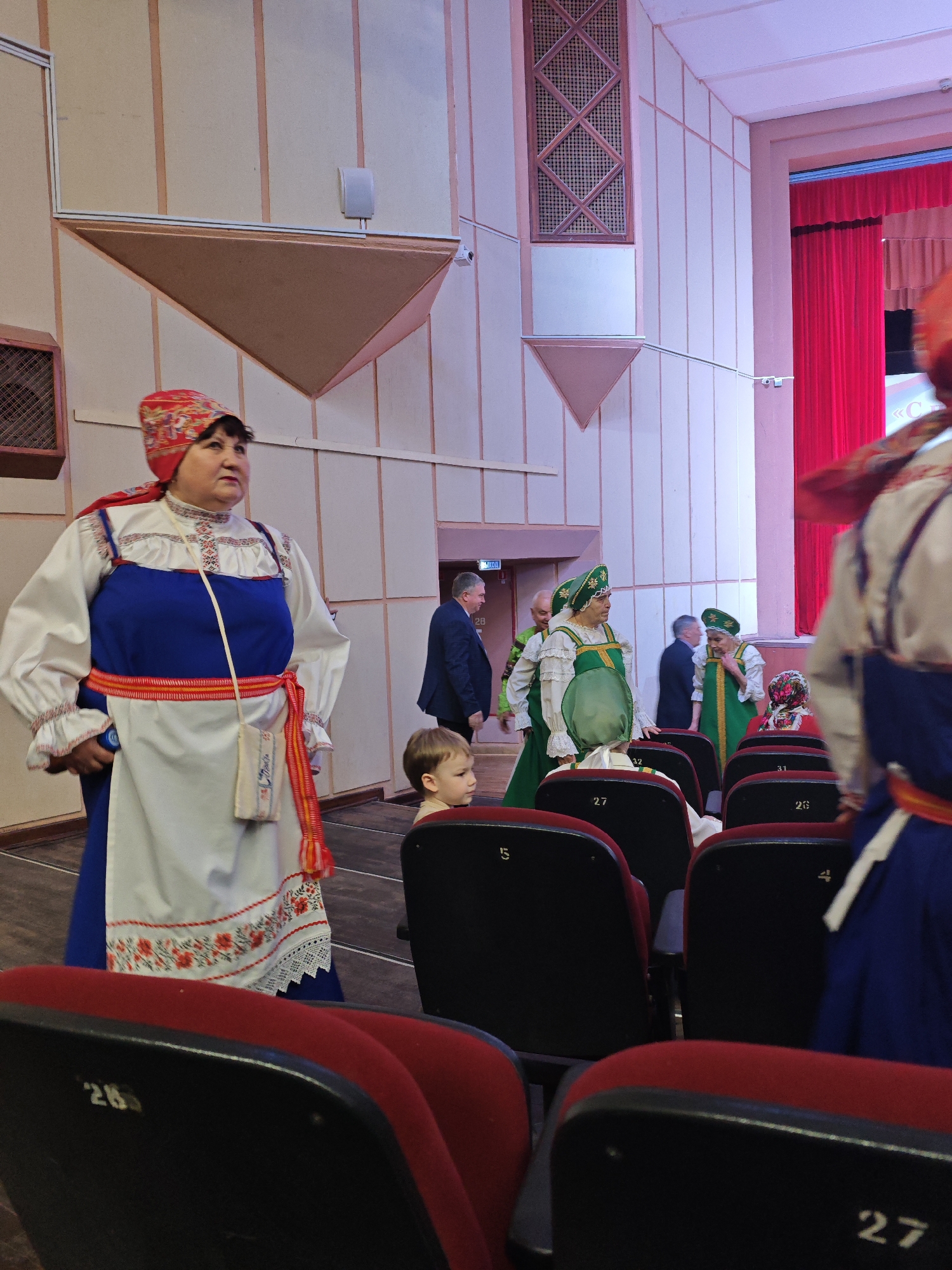 Captured indoors in what appears to be a theater or auditorium in Марковский, Russia, the image features a variety of people and objects.
The foreground showcases rows of unoccupied, dark red, cushioned seating with numerical labels. In the middle ground, a group of individuals dressed in traditional folk costumes are present. One woman, clad in a blue and white outfit with a red patterned headscarf, stands out prominently. She is flanked by others, some wearing green attire. Several men in business attire are visible in the background, near an exit door with a green sign. A young child is seated among the rows of seating.
The background reveals parts of the stage with red curtains. The walls of the room are light in color, featuring rectangular and triangular architectural elements. The scene conveys a cultural event or performance taking place in a public space.