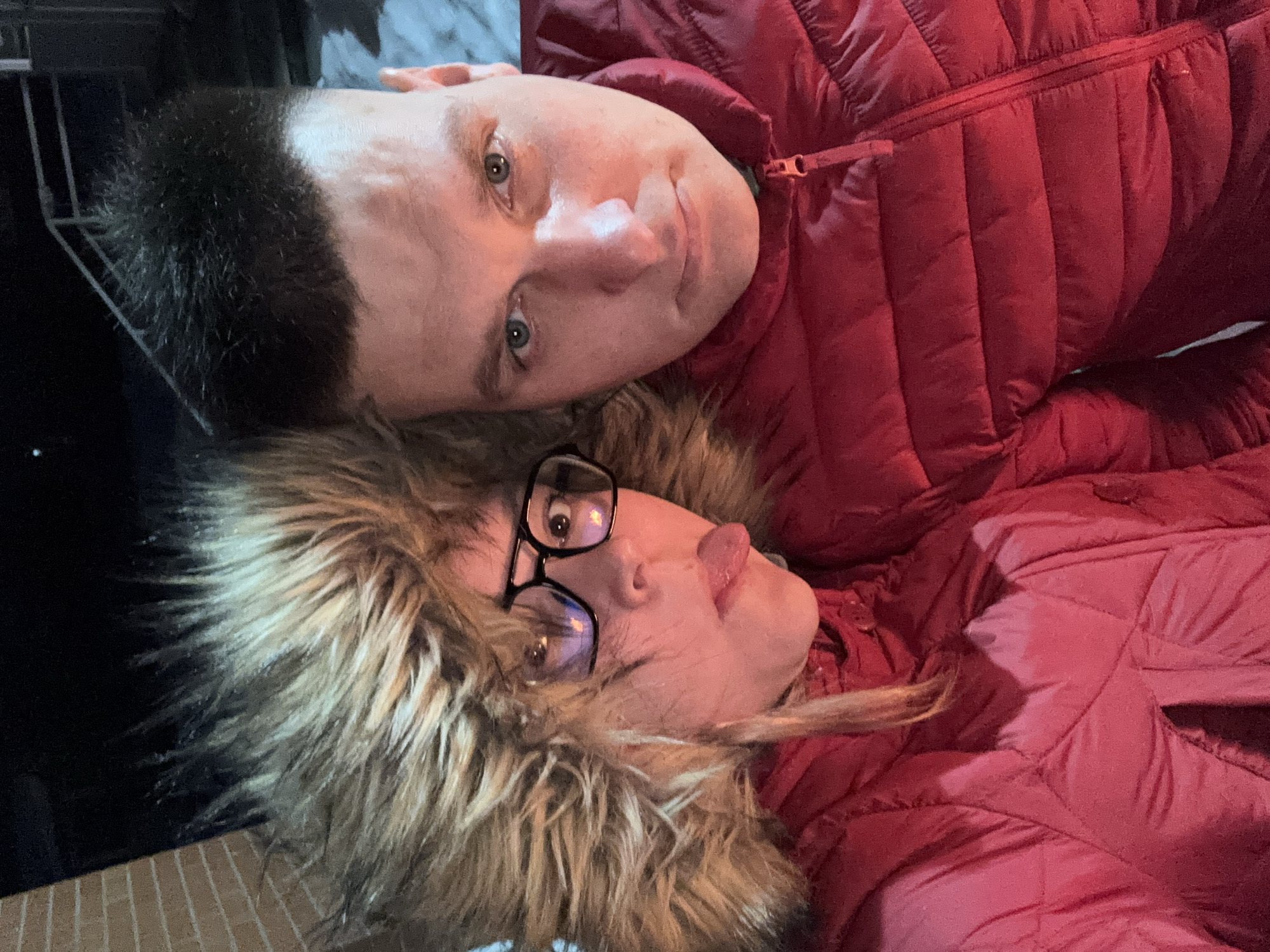A close-up selfie captured outdoors at night in Русский Вожой, Russia, features two individuals. On the left, a female wears a dark red padded winter jacket with a prominent light brown fur-lined hood, black-rimmed glasses, and has her tongue playfully extended. On the right, a male wears a matching dark red padded winter jacket and possesses short, dark, spiky hair with a neutral expression, looking directly at the camera. A partial view of a light brown brick wall is visible in the upper left background, while the upper right background is dark with hints of a white surface, possibly snow, consistent with a cold, nighttime setting.