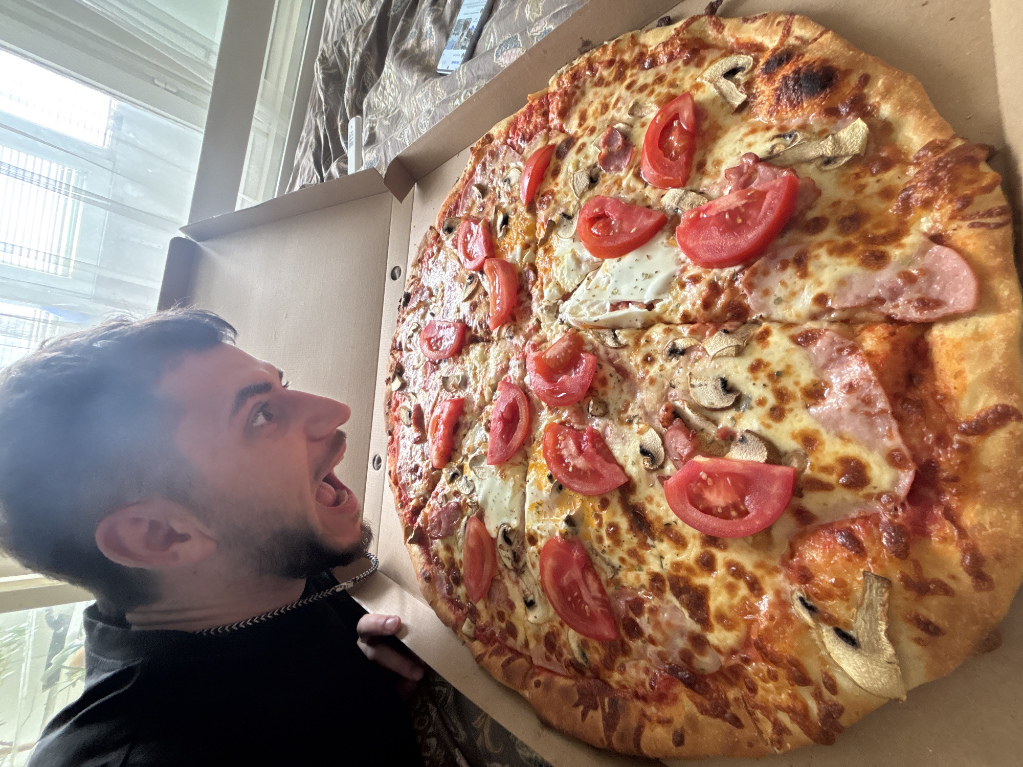 A man is shown looking at a pizza in a pizza box. The pizza is very large and has tomatoes, cheese, mushrooms, and ham on it. The man has dark hair and a beard and is wearing a black shirt. The pizza is in a cardboard box. A window and brown patterned curtain are in the background. The location is unknown.