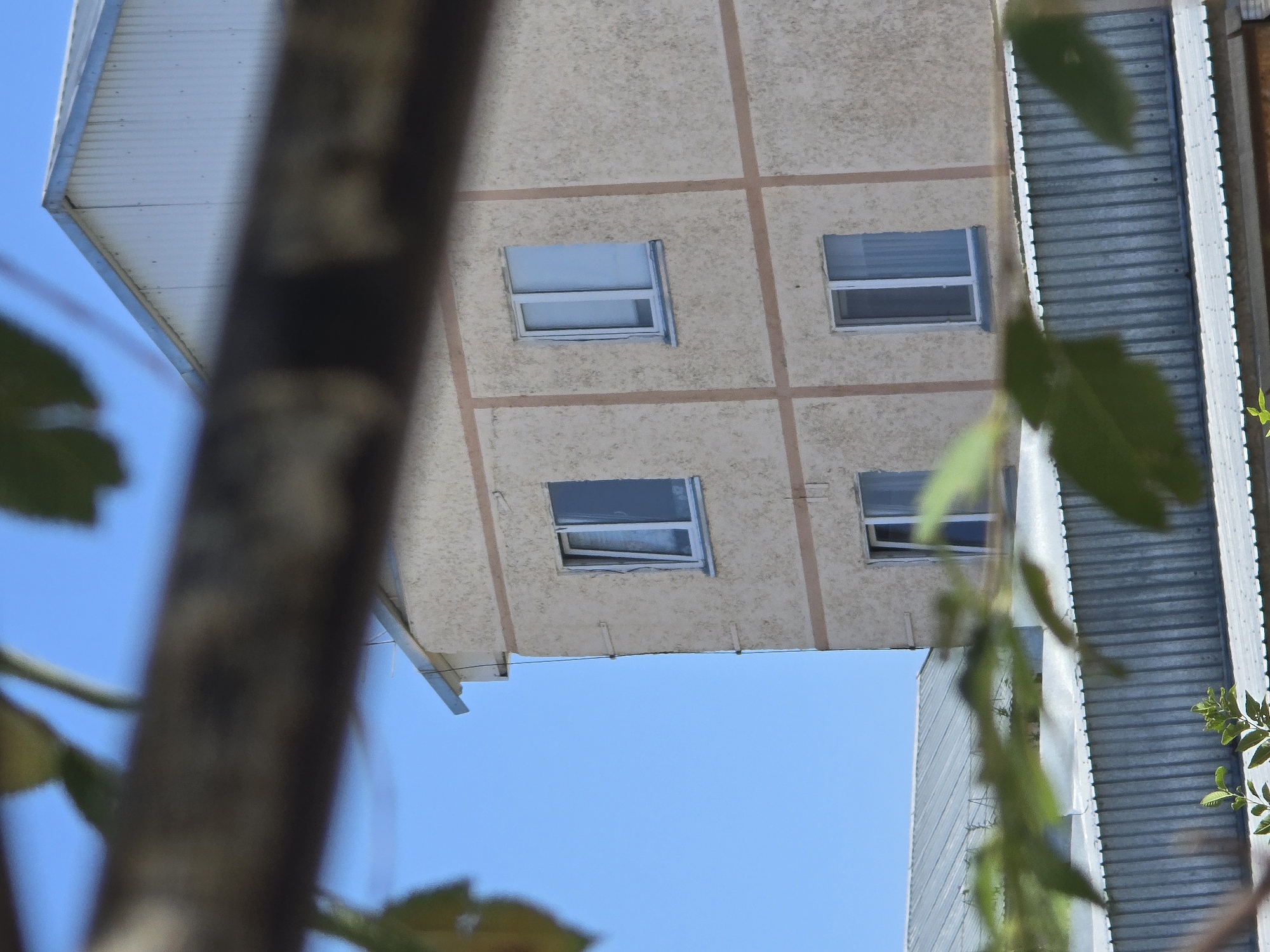 The image shows a beige building with a white roof and blue sky in Qaratau, Kazakhstan. The building has several windows, some of which are open. A corrugated metal structure is visible in the foreground, partially obscuring the view of the building. Green leaves are also visible in the foreground, suggesting that the photo was taken outdoors. The overall scene appears to be a residential area.