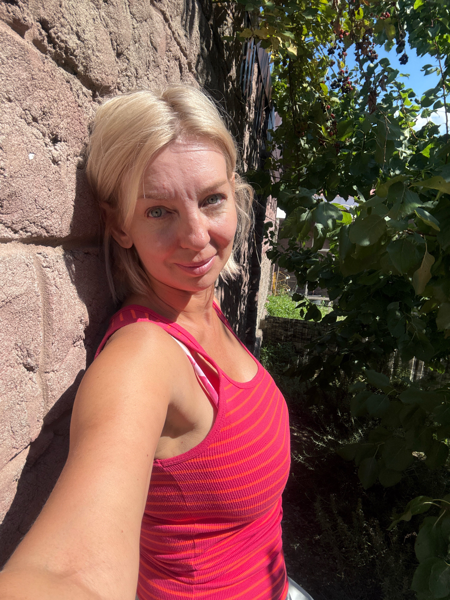 In Abrau-Dyurso, Russia, a blonde woman with blue eyes stands leaning against a textured, pinkish-brown stone wall. She is wearing a pink ribbed tank top with orange stripes. Behind her, a tree with dark green leaves and red berries is visible, with a clear blue sky above. The scene is illuminated by bright sunlight, casting shadows on the wall and the woman.