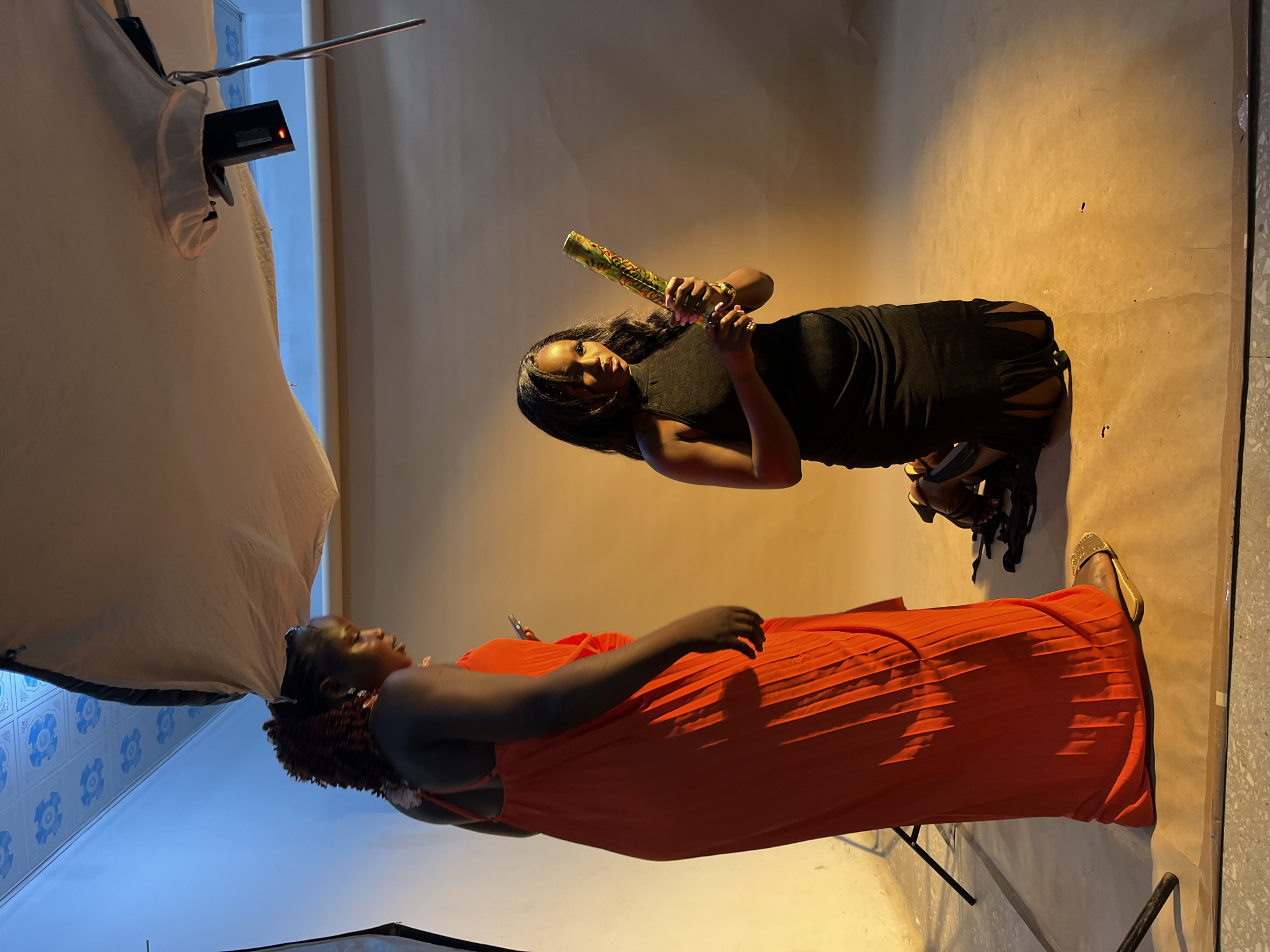 The image depicts an indoor studio setup with two Black women. The woman on the left stands wearing a floor-length orange dress and holds a smartphone. The woman on the right kneels, wearing a black sleeveless dress and holding a confetti popper. The background is a seamless paper backdrop, illuminated by warm lighting, creating an orange-brown hue. A light diffuser is positioned overhead, and a flash unit is visible. The floor appears to be concrete. The scene is consistent with a professional photo shoot. The location is not identifiable as Gura Top, Nigeria from the visual information provided.
