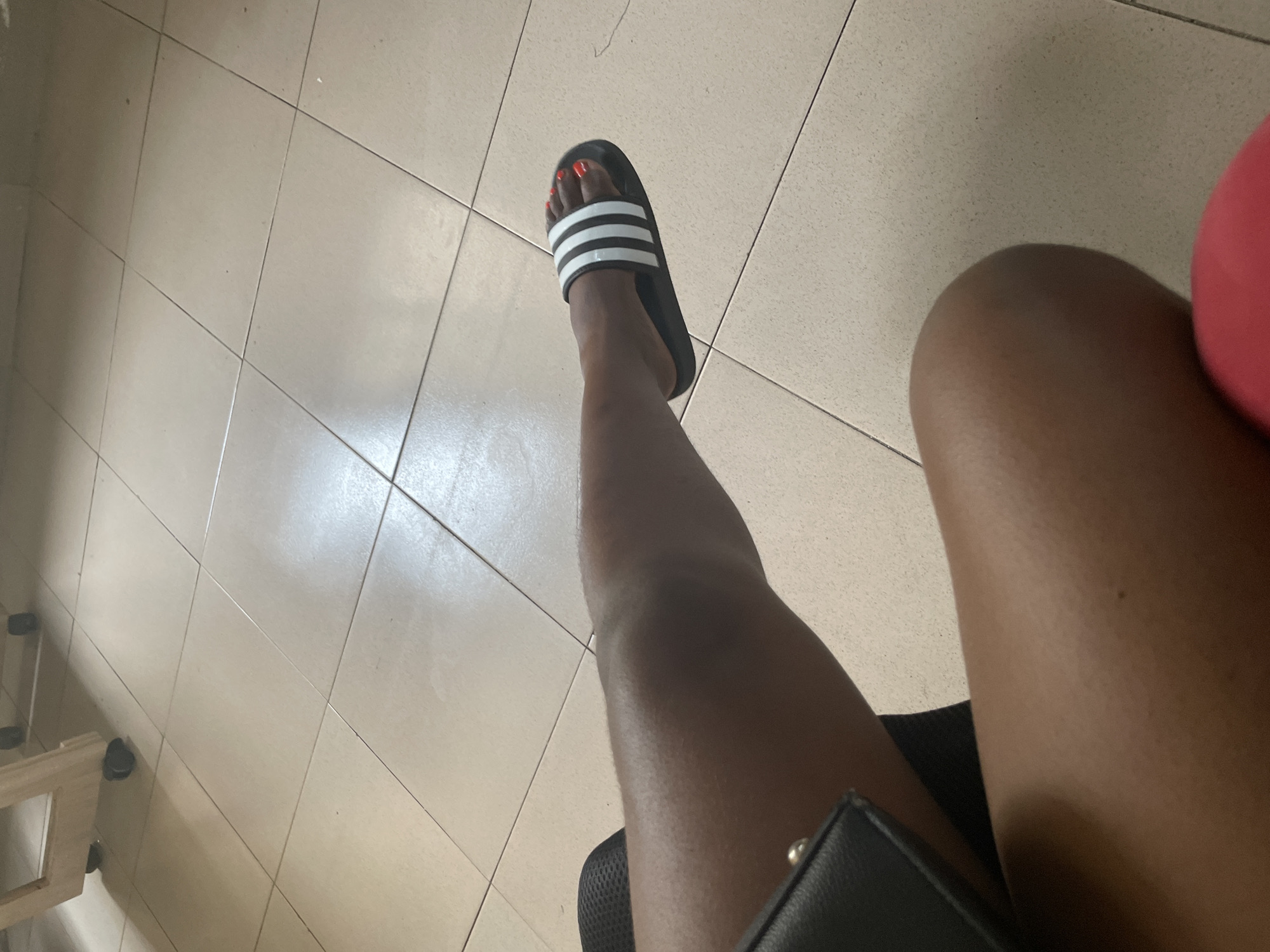 A medium shot shows a person's legs and feet from a seated perspective. The person is wearing black Adidas slides with white stripes on their feet, and their toenails are painted a bright orange. The individual's skin tone is dark. The legs are positioned on a tiled floor with a light beige grout. To the left, a portion of a black purse is visible. In the background, a light brown wooden structure with wheels is partially visible. The scene appears to be indoors, possibly in a waiting area or living space in Ado, Nigeria.
