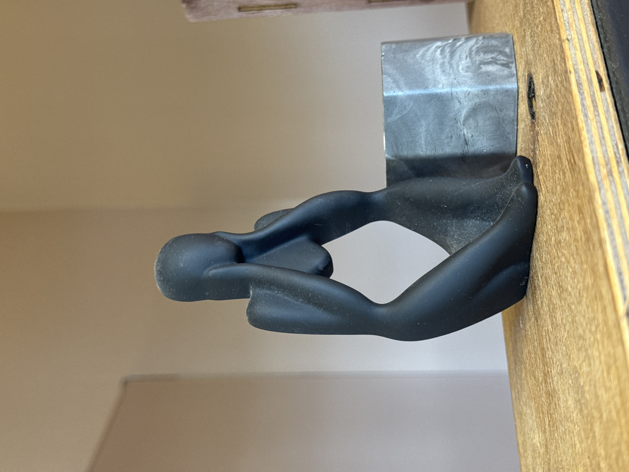 This is a close-up, vertically oriented, eye-level shot of a black, abstract sculpture resembling a human figure in a seated or reclining pose. The sculpture is made of a matte, dark material. It is attached to a light-colored wooden surface, which has visible grain and some imperfections. To the left of the sculpture, a metallic object with a brushed or textured surface is also visible, seemingly part of the mounting or display. The background is a plain, neutral-colored wall, out of focus. The lighting is soft and appears to be from an indoor source. There is no visible text in the image. The scene appears to be a still life or an indoor display, possibly in a studio or a home setting. The orientation of the image suggests it might have been taken with a phone camera.