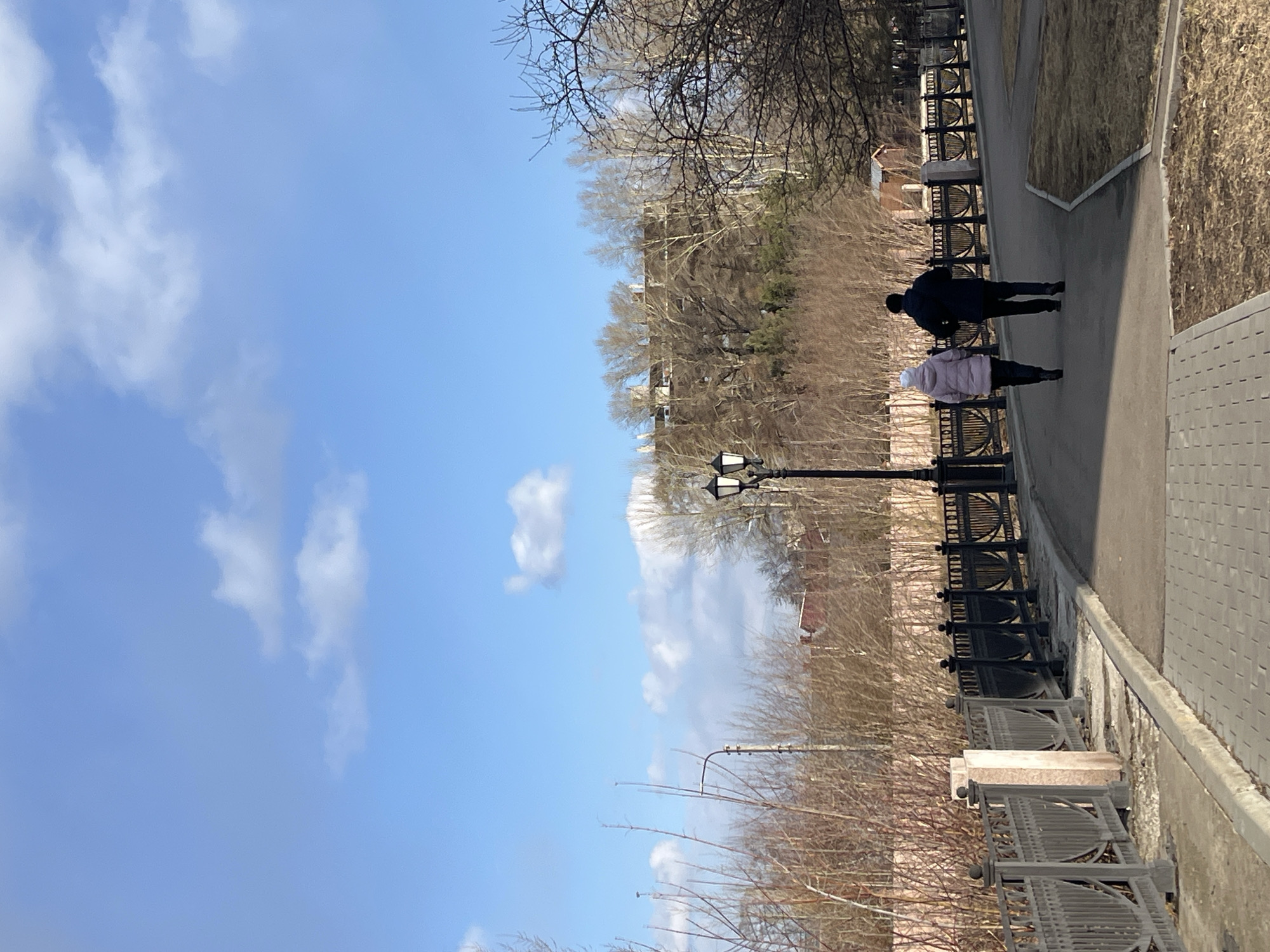 A photograph shows a sidewalk in Krasnoyarsk, Russia, along which two people are walking. One person wears a dark jacket, and the other wears a light pink jacket and a white hat. A black iron fence separates the sidewalk from a brown grassy area. A black lamppost stands along the fence. Beyond the fence, leafless trees are visible. A blue sky with scattered clouds is above. The sidewalk is gray and appears to be made of concrete. The image is rotated 90 degrees counterclockwise.