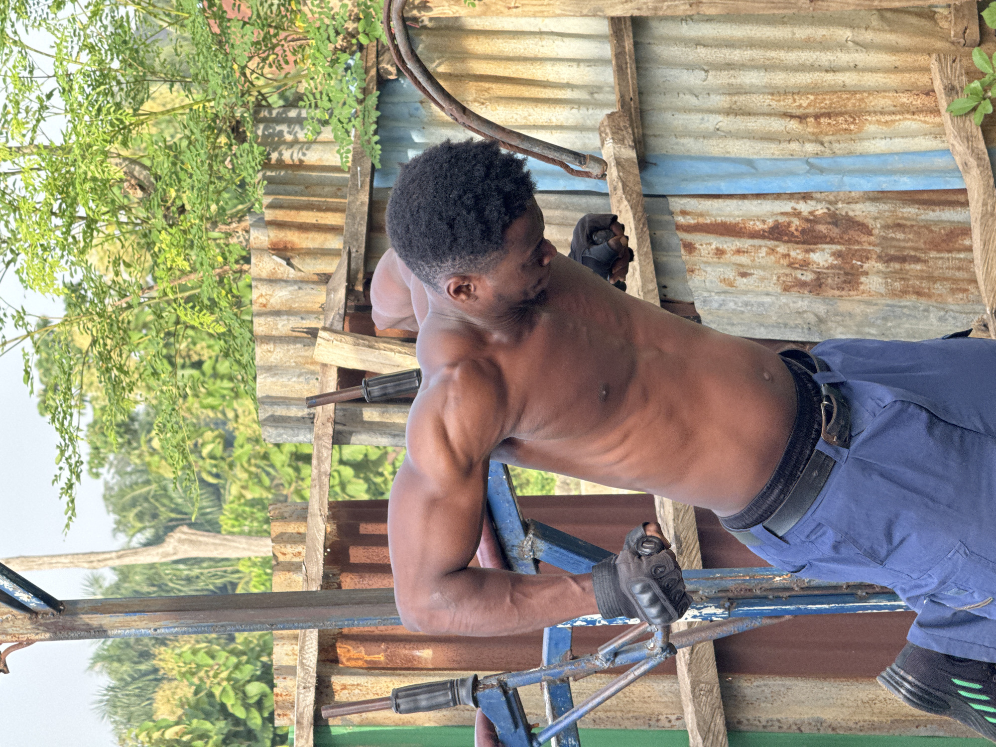 The image depicts a shirtless man with muscular arms and a dark complexion performing dips on a homemade weight apparatus. He is wearing black workout gloves and blue cargo pants with a black belt. The background consists of corrugated metal fencing, wooden planks, and lush green vegetation, suggesting an outdoor setting in a rural or semi-rural environment. The overall impression is one of a functional, perhaps improvised, outdoor gym. The image does not contain any identifiable landmarks or features that would specifically place it in Akwanga, Nigeria.