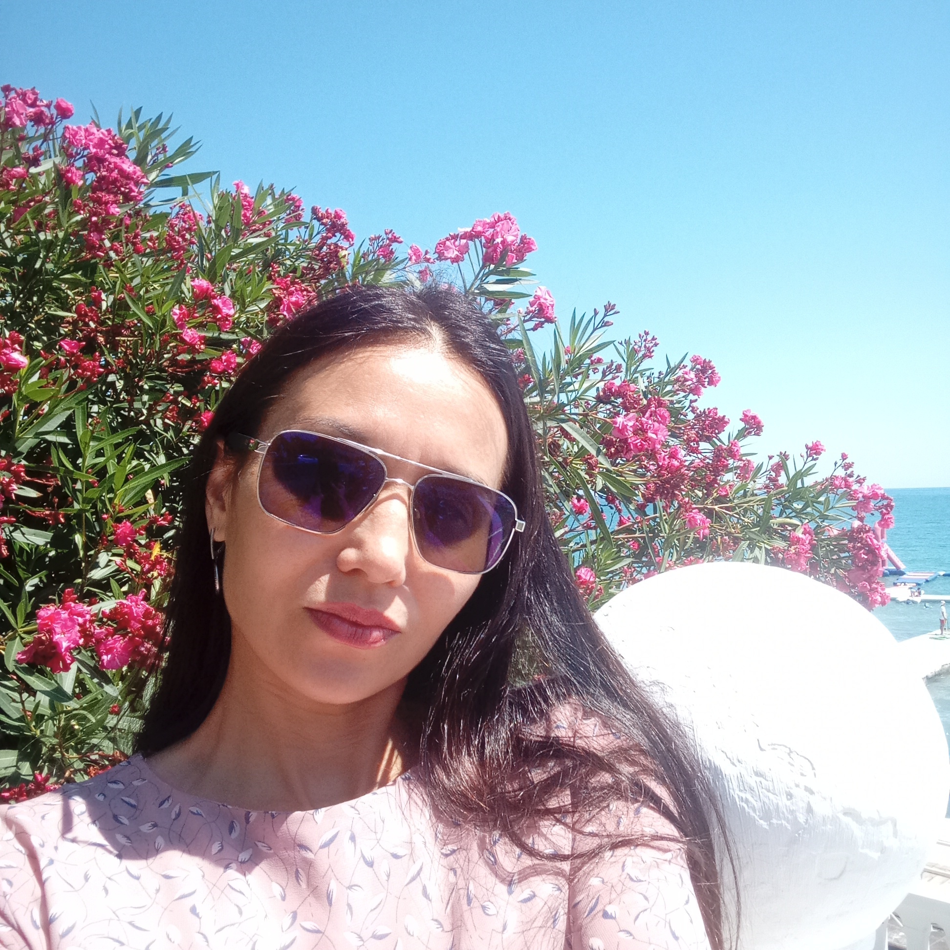 A woman is taking a selfie outdoors, with a clear blue sky and the sea visible in the background. She has long dark hair and is wearing sunglasses and a light pink patterned dress. She is standing in front of a bush with bright pink flowers. To the right of the woman, there is a large white rounded object, possibly a sculpture or architectural element. In the distance, on the sea, there are some inflatable water attractions and a few people can be seen. The scene suggests a sunny day in a coastal resort. The specific location is not identifiable as Sochi, Russia, based solely on the visual information present.