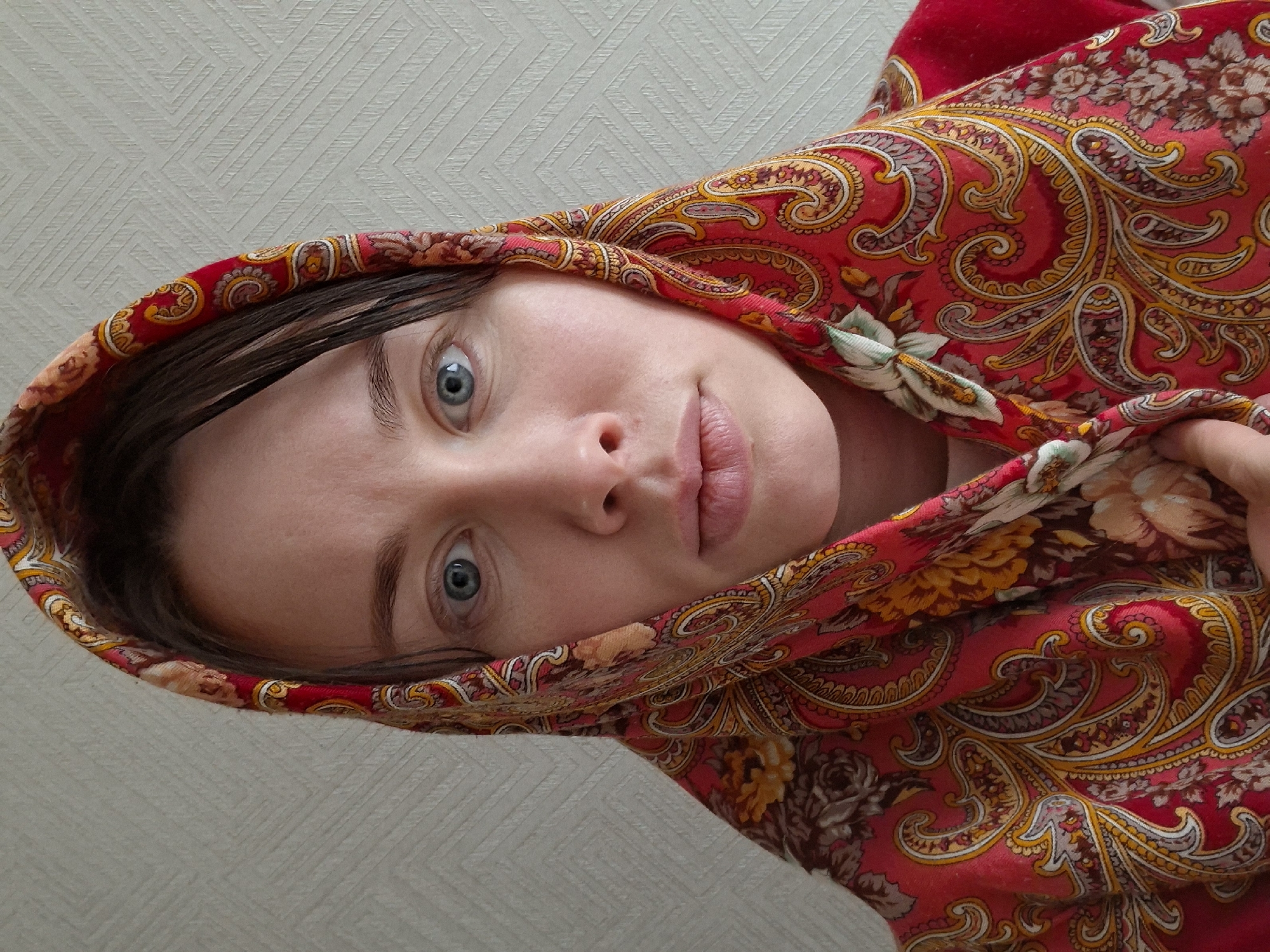 A close-up portrait features a person, likely a woman, looking directly at the camera with wide, clear blue eyes. She has fair skin and visible brown hair peeking out from under a headscarf. Her lips are slightly parted, and her expression is neutral.
The dominant feature in the image is a vibrant red headscarf or shawl worn by the subject. The fabric is adorned with an intricate and colorful pattern, primarily featuring large paisley motifs and floral elements in shades of red, yellow, orange, white, and muted brown/grey. The headscarf wraps around her head, framing her face and covering her hair.
The background consists of a light-colored wall with a subtle, repeating geometric textured pattern, suggesting an indoor setting. The image is rotated 90 degrees clockwise from a typical vertical orientation, giving the impression of the subject looking upwards or having her head tilted significantly.
No specific time of day or weather conditions can be discerned from the indoor setting. There are no identifiable location cues beyond the generic wall texture. No text is visible in the media file.