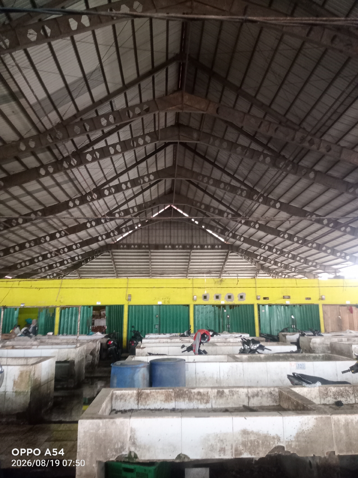 The image shows the interior of a covered market in Jambi City, Indonesia. The market has a high, corrugated metal roof supported by metal beams and trusses. The walls appear to be a combination of yellow painted sections and green corrugated metal roll-up doors or partitions. Below the roofline, there are numerous stalls or market stands constructed from concrete. Some of these stands contain blue plastic barrels. Several motorcycles are parked within the market area, and some appear to be on or near the market stands. A few individuals are visible in the background, near the green partitions, though their activities are not clearly discernible. The ground appears to be wet or damp in places, suggesting it may be a fish market or a location where water is frequently used. The overall scene depicts a functional, potentially busy, indoor market environment.
