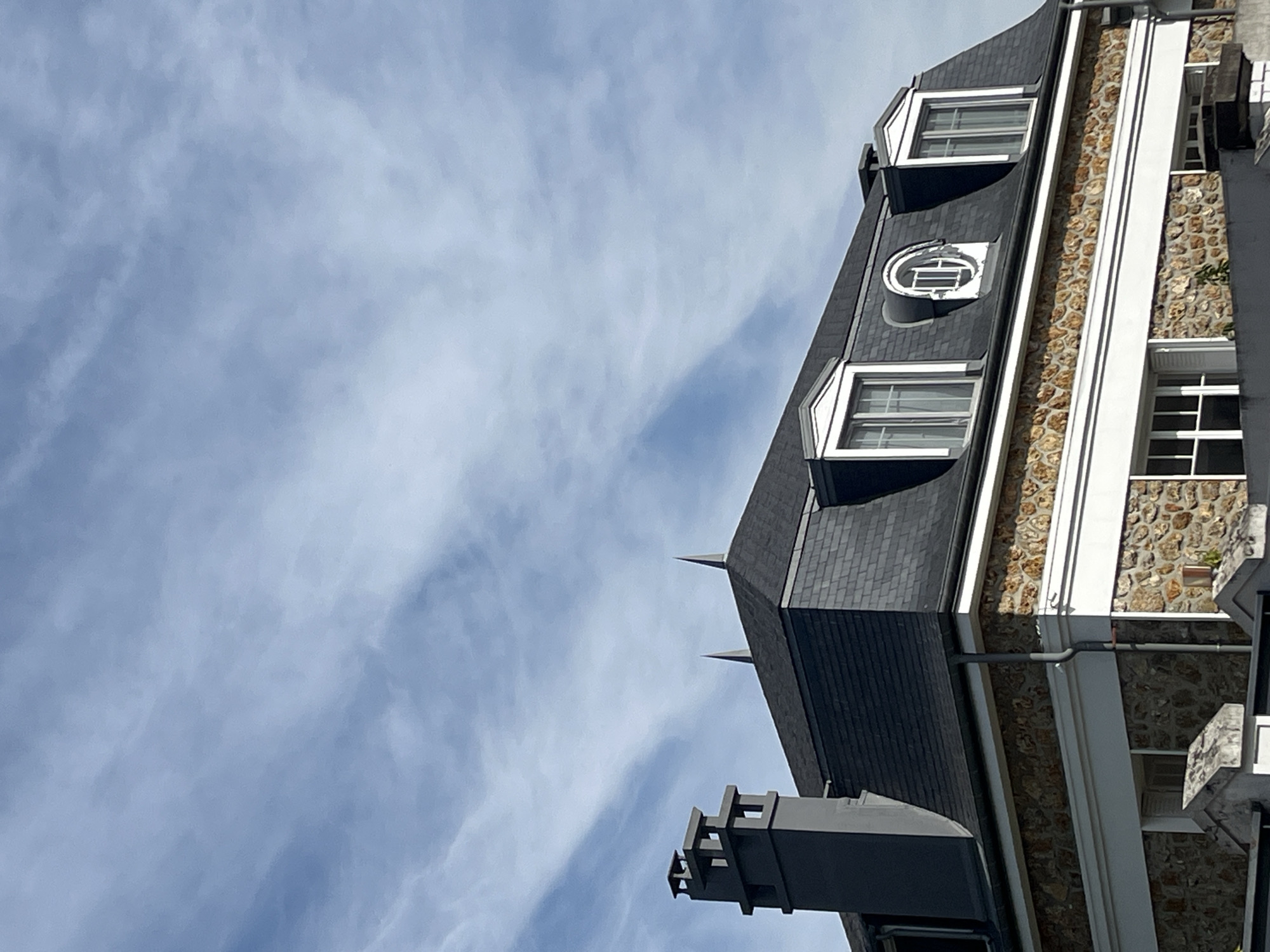 The image shows the roof and upper facade of a stone building under a cloudy blue sky. A dark grey slate mansard roof with two peaked dormer windows and one circular dormer window is prominent. A tall, grey, stepped chimney is visible on the left side of the roof. The building's facade is constructed of rough, irregularly shaped stones in shades of brown and orange, bordered by a white decorative band. Below this, more windows are partially visible. The scene appears to be an exterior view of residential architecture. Based on available information, this building is located in Chevilly-Larue, France.