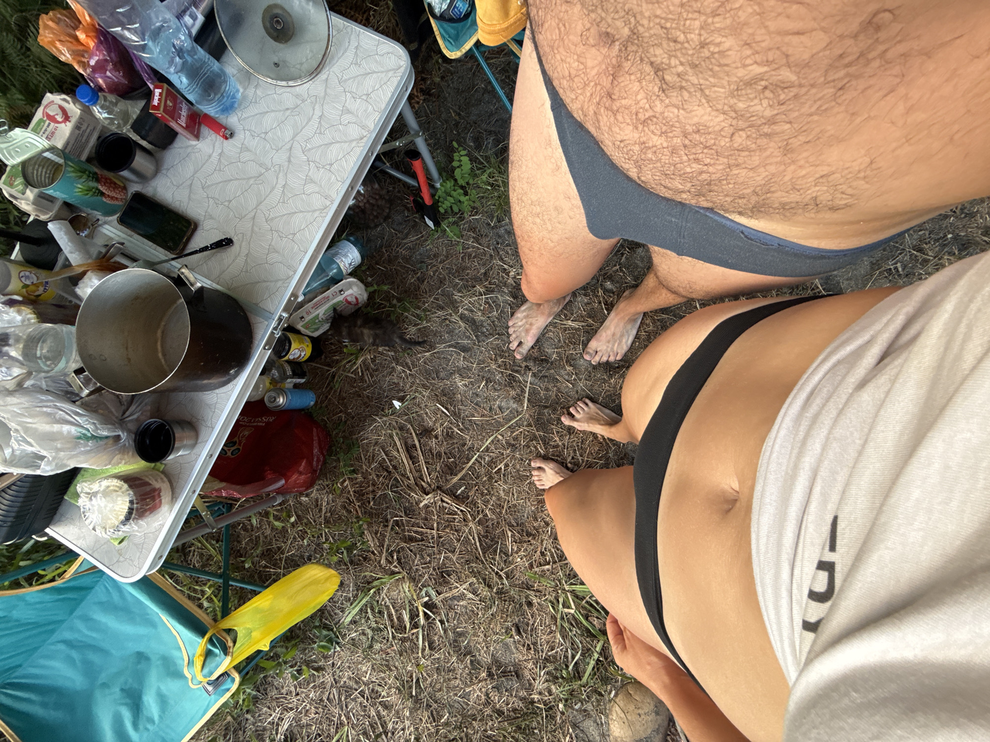 This media file shows a close-up perspective of two individuals from the waist down, outdoors in a rustic setting. The scene appears to be a campsite, with a small folding table laden with various items like a pot, cans, bottles, a pack of cigarettes (Marlboro), a smartphone, and plastic bags. One individual, whose legs are visible in the upper right of the frame, is wearing dark briefs and has a significant amount of body hair on their thighs and abdomen. Their feet are on the dry, grassy ground. The second individual, in the lower portion of the frame, is wearing black underwear and a white t-shirt that is pulled up, revealing their midriff. Their legs are also on the ground, and their feet are visible. The ground is covered in dry grass and pine needles, suggesting a natural, possibly wooded, environment. In the background, a camping chair with a teal-colored fabric seat is partially visible, as is a yellow plastic bag. The lighting suggests it is daytime, with the sun casting shadows. The overall mood is casual and relaxed, hinting at a camping or outdoor recreational activity. There is no discernible text other than that on the cigarette pack. The location is identified as Yuada-Gulrypsh, Abkhazia.