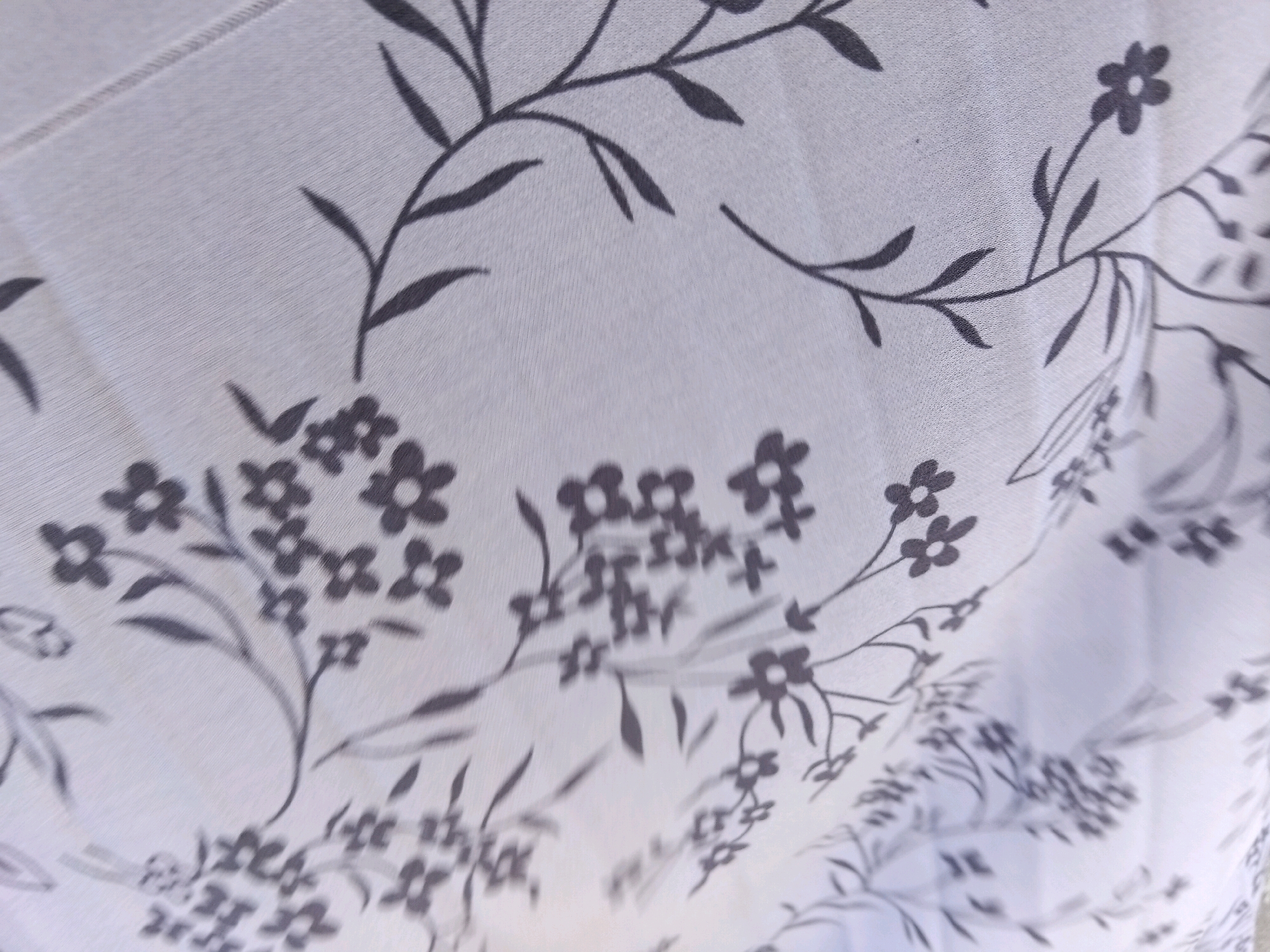The image displays a close-up, slightly out-of-focus view of a light-colored fabric with a delicate, dark gray floral pattern. The pattern consists of thin, branching stems with small leaves and clusters of simple, four-petaled flowers. The fabric appears to be soft and possibly sheer, as indicated by the way it drapes and wrinkles, creating subtle shadows and highlights. The overall impression is one of a light, airy textile, likely used for curtains or apparel. There is no indication of the city of Kepuh Kiriman, Indonesia, or any specific environmental context within the image.
